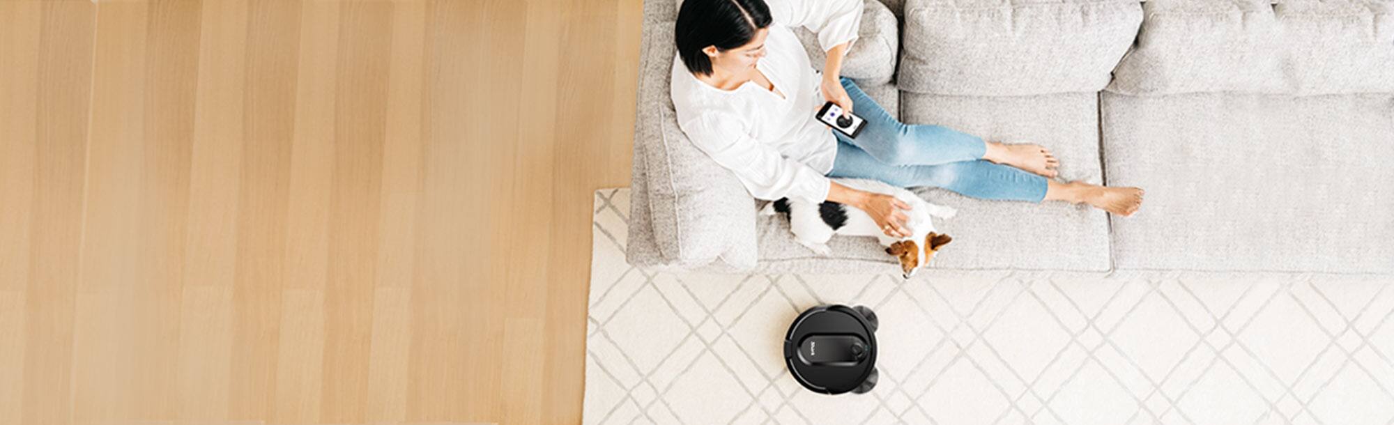 Person on sofa with a dog and a smartphone that is controlling a robot vacuum on carpet below