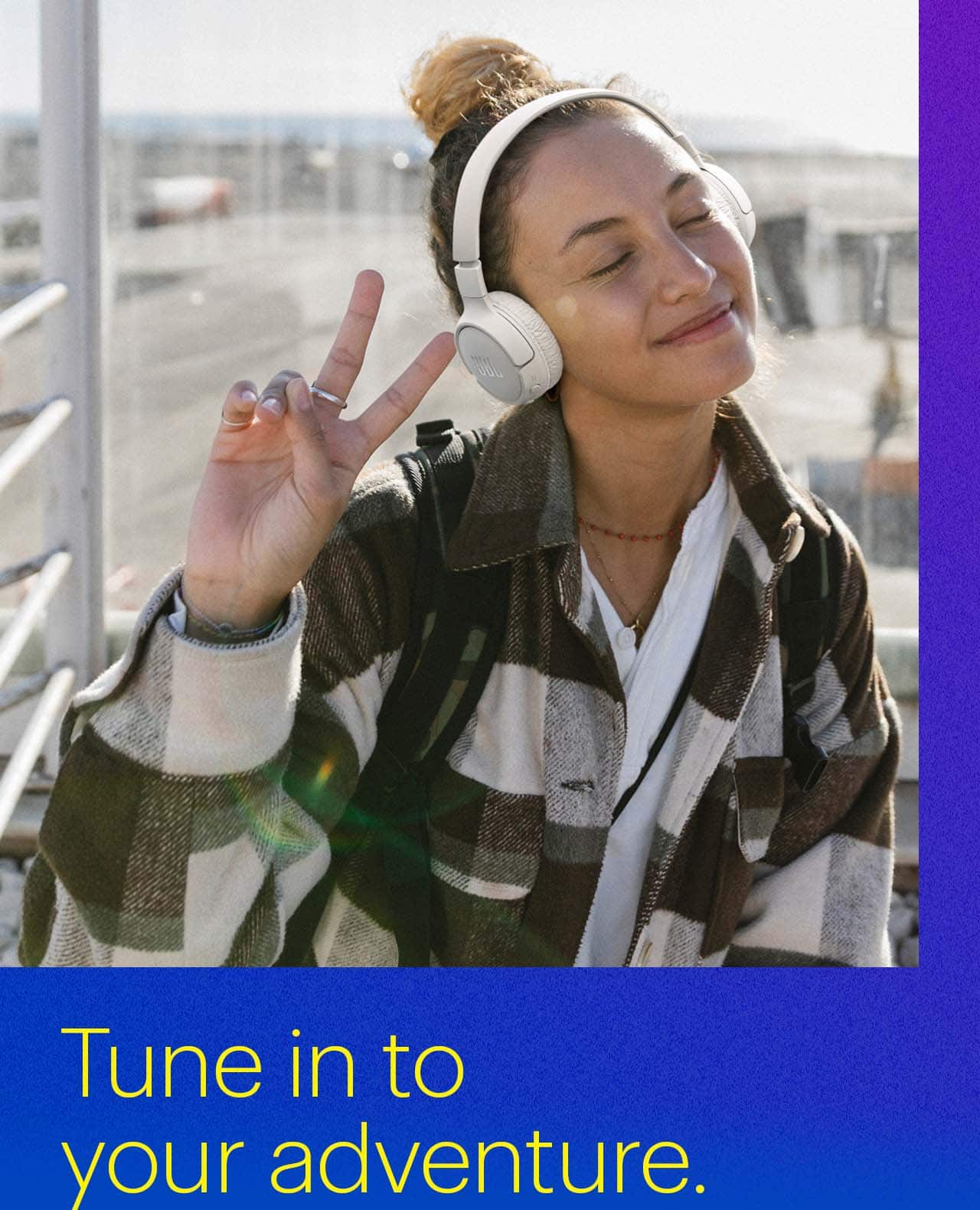 Tune in to your adventure.  Person smiling wearing JBL Adaptive Noise Cancelling Wireless Headphones at airport.