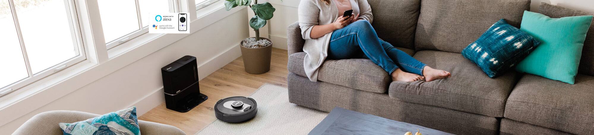 Person sitting on couch with robot vacuum on floor next to docking station. Works with Alexa. Works with the Google Assistant.