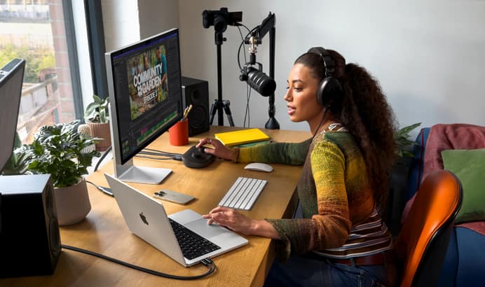 A woman in her home working at her desk with MacBook Pro connected to two external displays, wearing headphones and speaking into a microphone