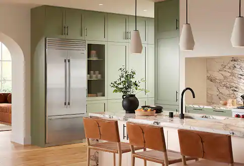 Stainless steel refrigerator, green cupboards, white countertops and island with brown chairs