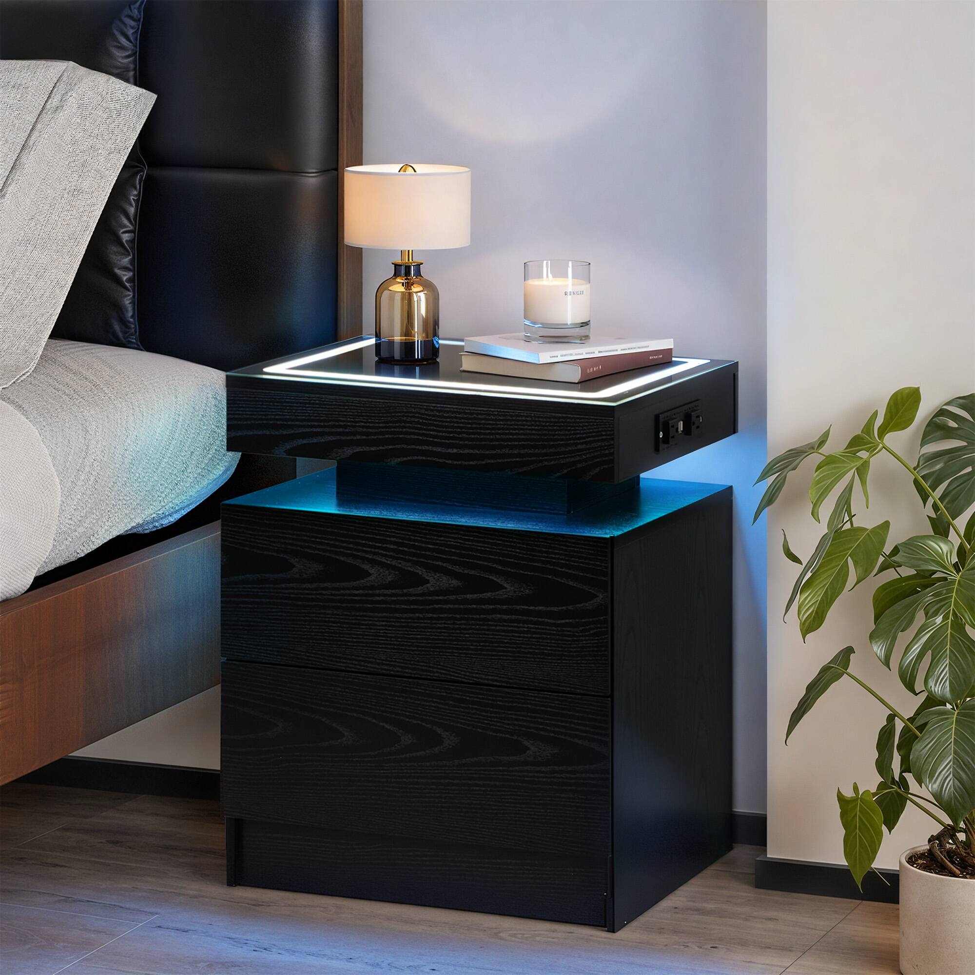 The image shows a modern bedroom nightstand setup. Here's a description based on the image:

- A black leather headboard with a gray pillow.
- A black nightstand with a glass top and blue LED lighting underneath.
- On the nightstand:
  - A brass lamp with a white shade.
  - A white candle in a glass holder.
  - Two books stacked on top of each other.
- A potted plant with green leaves is placed on the floor next to the nightstand.
- The wall is painted in a light color, and the floor is wooden.

The text on the books is not clearly readable, but the setup is stylish and contemporary.