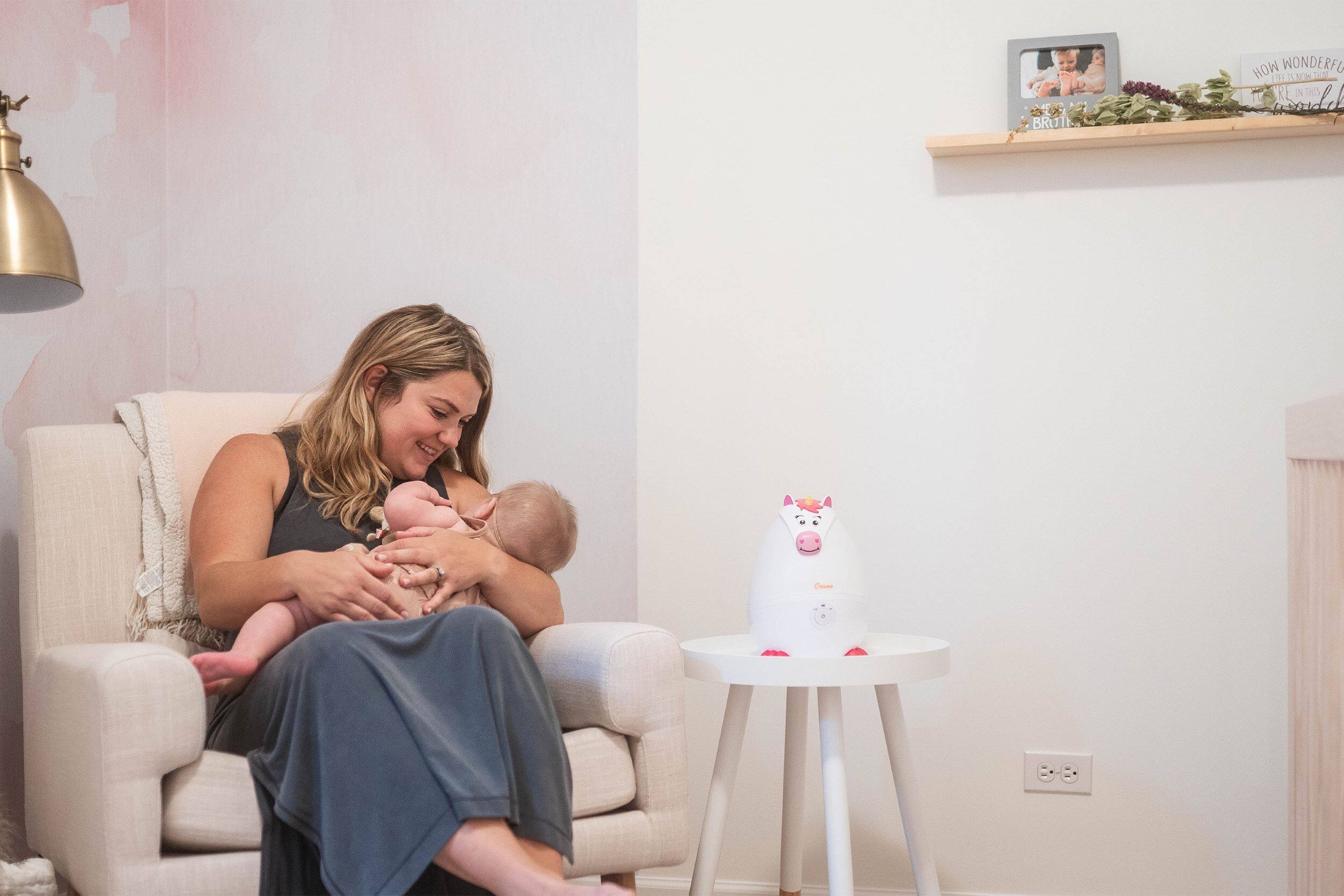 The image shows a woman sitting in a chair, holding a baby in her arms. The woman is smiling, and the baby is looking at her. The room has a white wall and a shelf on the wall. There is also a potted plant in the room. The woman is wearing a dress, and the baby is wearing a diaper. The scene appears to be a cozy and intimate moment between the woman and the baby.