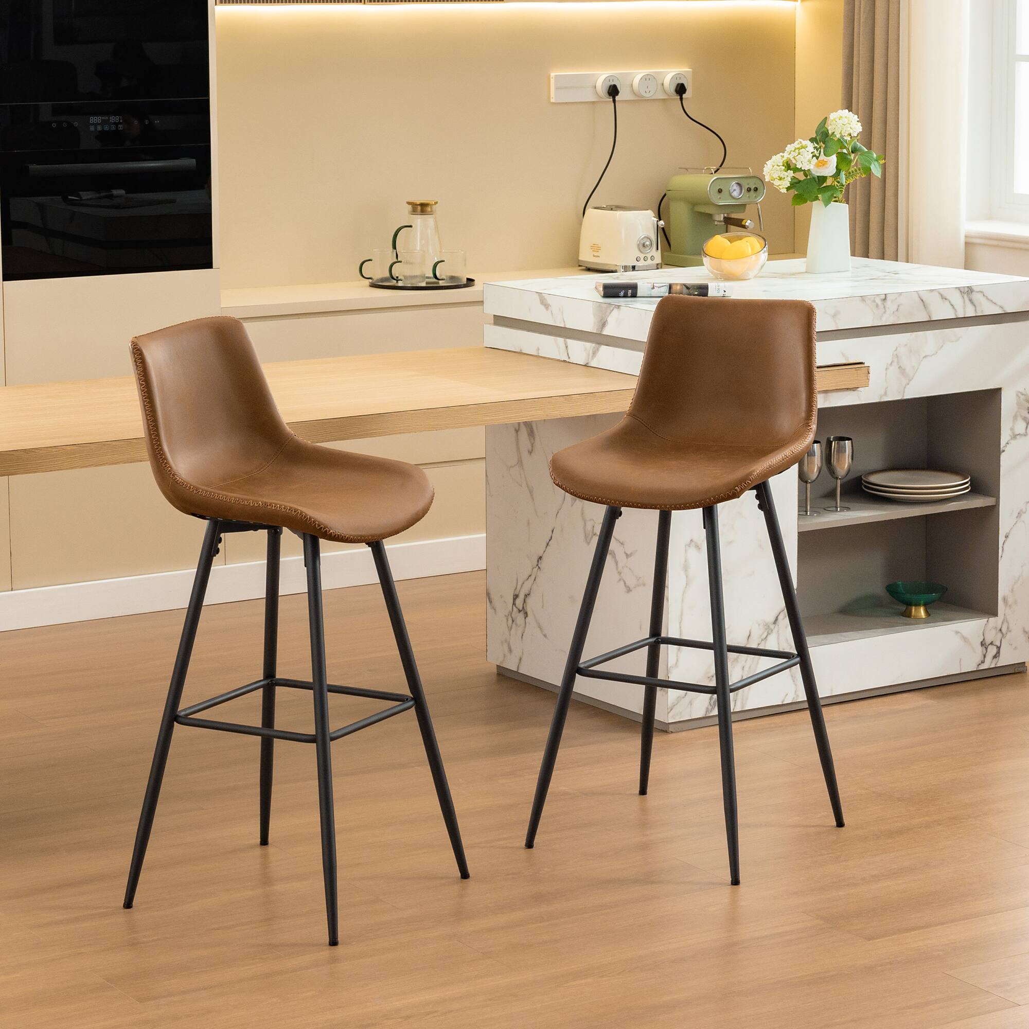 The image shows a modern kitchen with a marble island countertop. There are two brown leather bar stools with black metal legs in front of the island. On the countertop, there is a green stand mixer, a bowl of lemons, a vase with white flowers, and some books. The backsplash is illuminated with under-cabinet lighting. The floor is wooden, and there is a built-in oven on the left side.