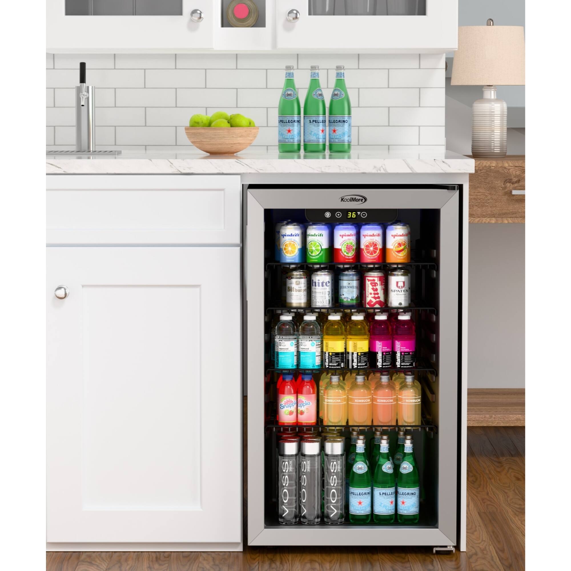 The image shows a refrigerator with a variety of drinks inside. There are several bottles of water, juice, and soda, as well as a bowl of fruit on the countertop. The refrigerator is filled with a wide assortment of drinks, including aloe vera, sparkling water, and other beverages. The scene suggests a well-stocked kitchen with a focus on healthy and refreshing options.