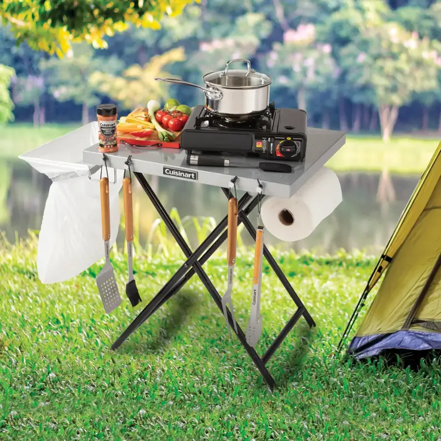 Cuisinart is a brand of cooking appliances and kitchen tools. In the image, there is a Cuisinart portable gas grill, which is a type of cooking appliance. The grill is placed on a table, along with other cooking utensils such as a spatula, tongs, and a knife. The table is set up in a park, creating an outdoor cooking experience.