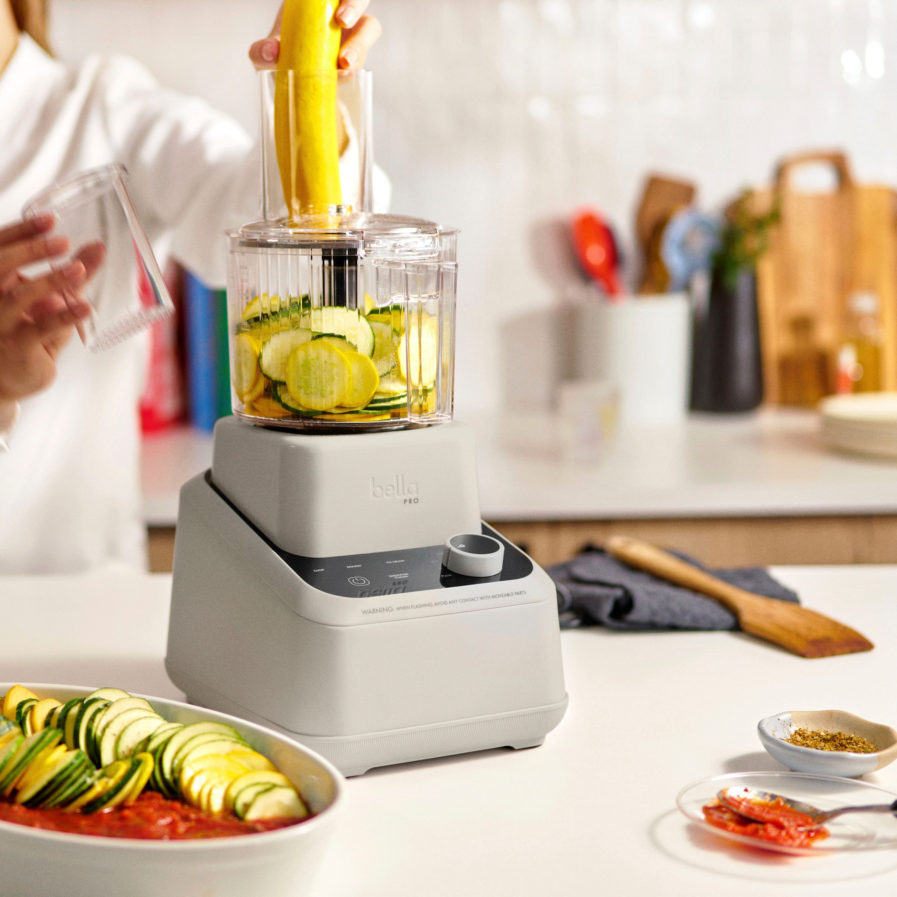 The image shows a person preparing food in a kitchen. They are using a blender to mix vegetables, including zucchini and cucumbers. The blender is placed on a countertop, and there are several books nearby. Additionally, there are bowls and a spoon on the countertop. The person is wearing a white shirt, and there is a knife in the scene as well.
