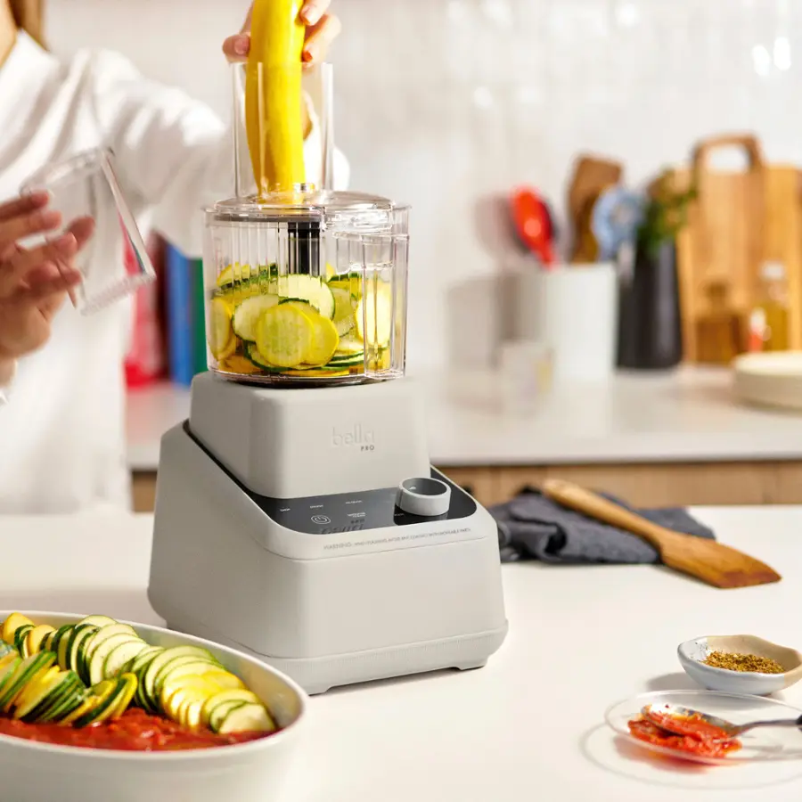 The image shows a person preparing food in a kitchen. They are using a blender to mix vegetables, including zucchini and cucumbers. The blender is placed on a countertop, and there are several books nearby. Additionally, there are bowls and a spoon on the countertop. The person is wearing a white shirt, and there is a knife in the scene as well.