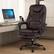The image shows a modern office setting featuring a dark brown leather office chair with a high backrest and armrests. The chair is positioned in front of a sleek, light-colored desk that holds a desktop computer with a large monitor displaying the Windows 10 desktop. The room has a minimalist design with vertical blinds covering the window, and the floor is made of light wood.