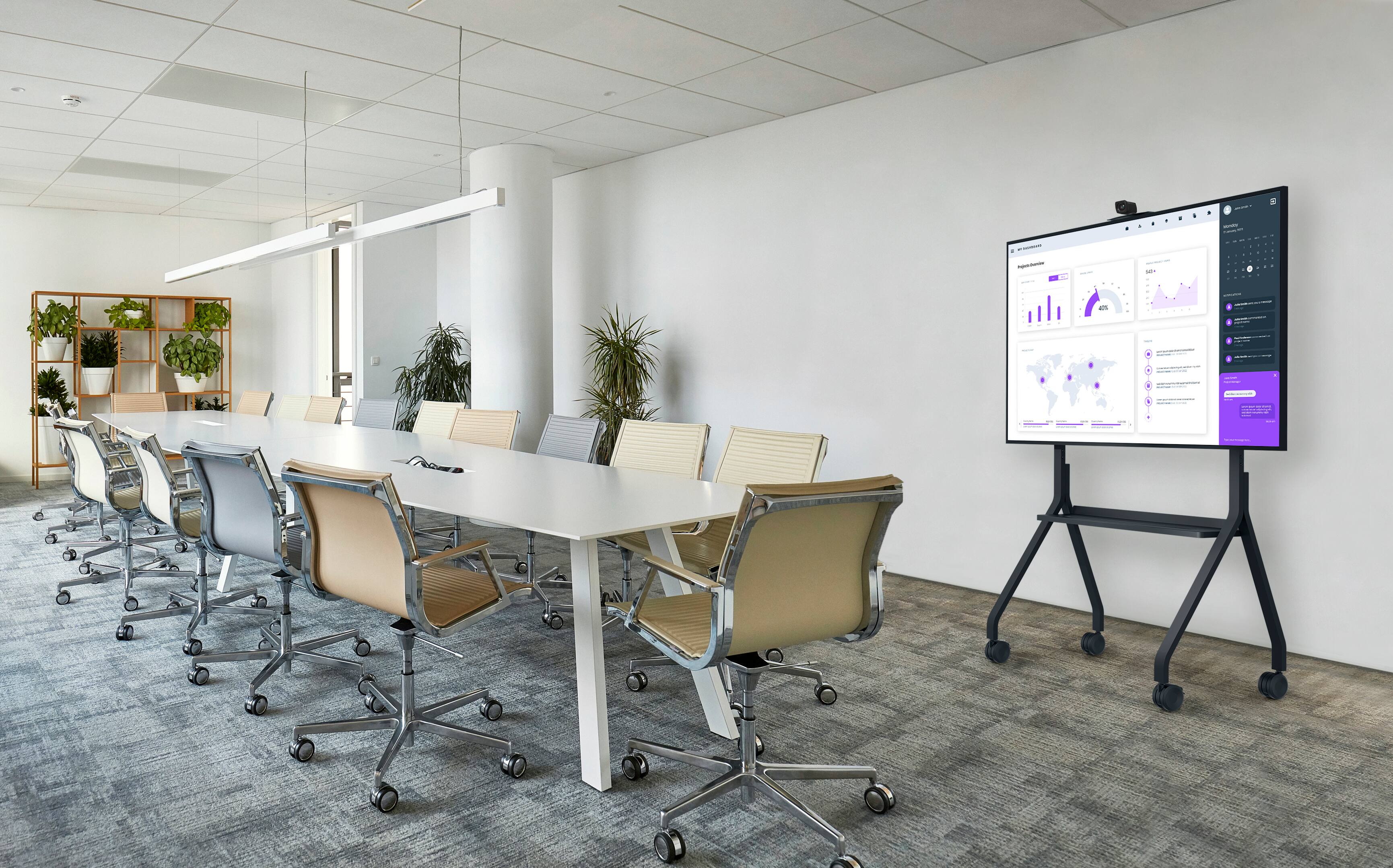 The image shows a conference room with a large screen TV mounted on the wall. The TV is displaying data and graphs, which suggests that it is being used for a presentation or meeting. The room is furnished with multiple chairs, some of which are arranged around the TV, and a dining table. There are also several potted plants placed throughout the room, adding a touch of greenery to the space. The overall atmosphere of the room appears to be professional and conducive to collaboration and discussion.