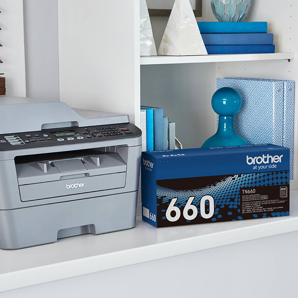 Brother 660 is a printer that is sitting next to a box of the same printer. The box is blue and has the name "Brother" on it. The printer is on a desk, and there are also some books nearby.
