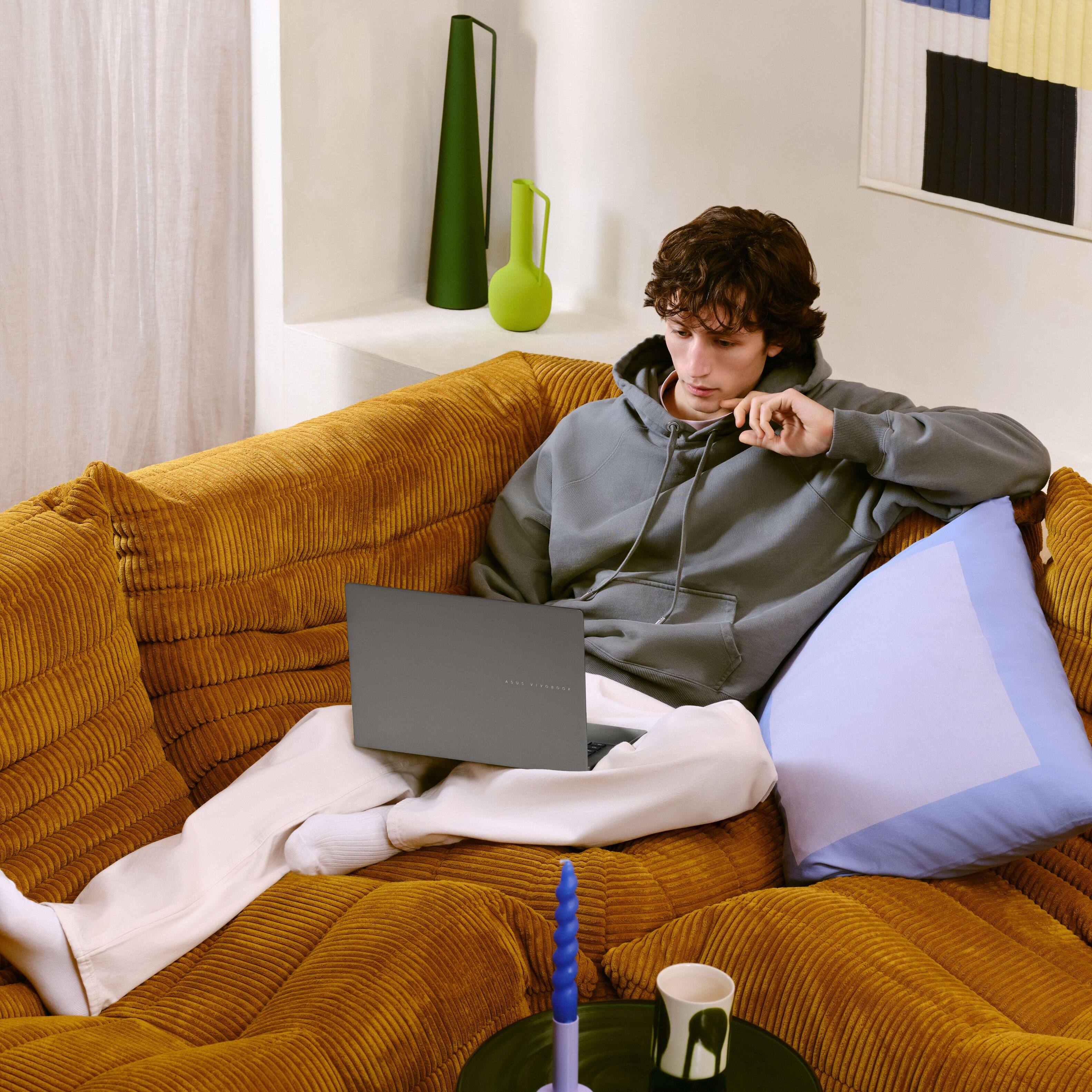 The image shows a person sitting on a mustard-colored couch, wearing a gray hoodie and white pants, using a laptop. The room has a modern decor with a green vase, a yellow pitcher, and a blue pillow.