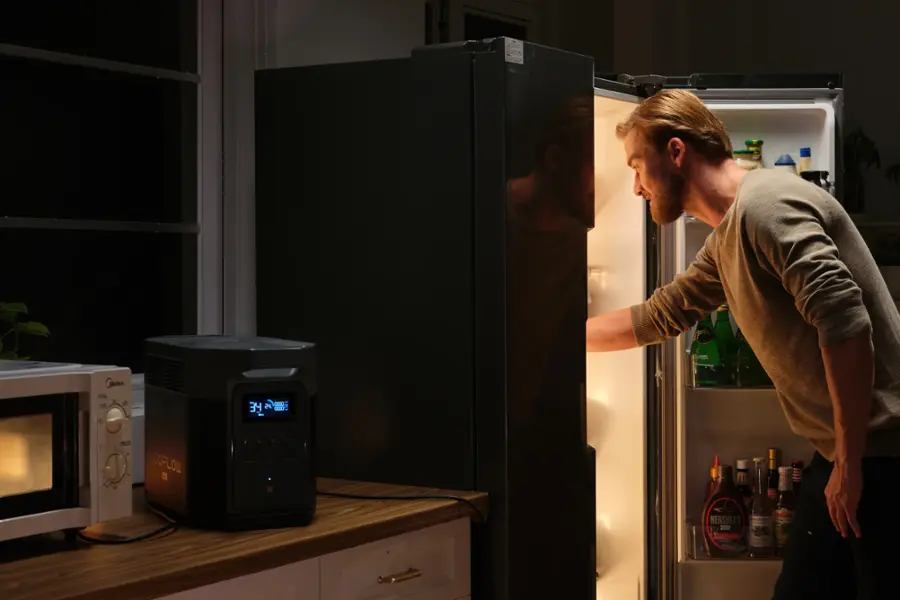 The man is standing in front of a refrigerator, which is open. The refrigerator is stocked with various items, including bottles of beer and soda. The man is likely looking for something to drink or eat. The scene takes place in a kitchen, with a microwave nearby.