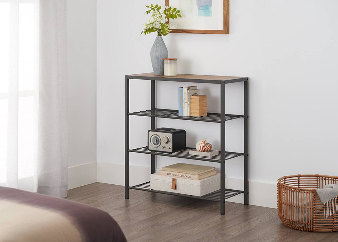 In the image, there is a bookshelf with a vase of flowers on top of it. The shelf is filled with various books, and there is a small basket on the floor next to the bookshelf. The scene appears to be in a bedroom, with a bed visible in the background.