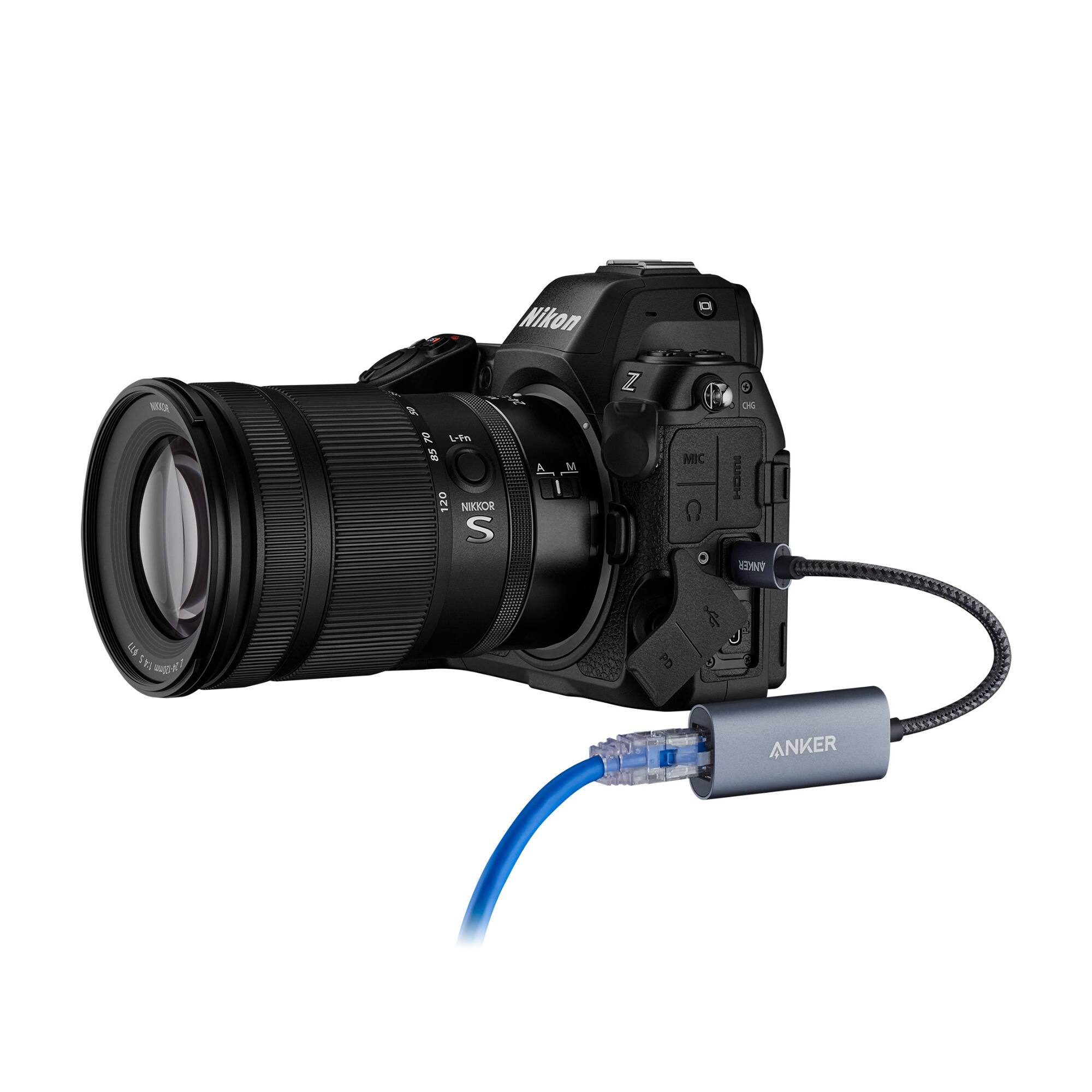 The image features a Nikon camera with a lens attached to it. The camera is connected to an Anker charger, which is plugged into the camera. The charger is likely used to power the camera or charge its battery.
