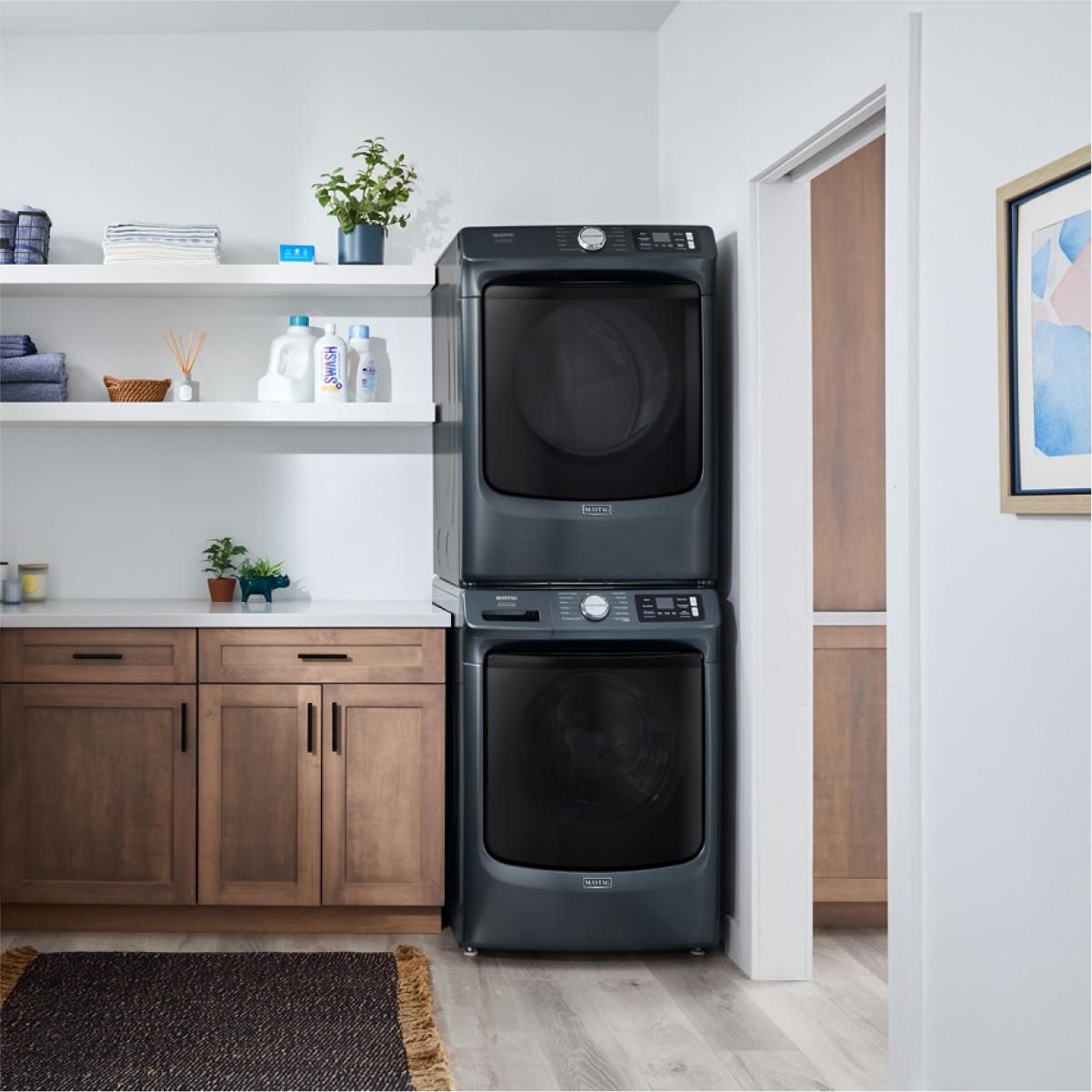 The image shows a laundry area with a stacked washer and dryer. On the shelves above the machines, there are towels, a plant, and laundry supplies, including a bottle labeled "SWASH." The cabinets below the shelves have a wooden finish, and there is a framed picture on the wall to the right. The floor is light wood, and there is a dark rug in front of the machines.