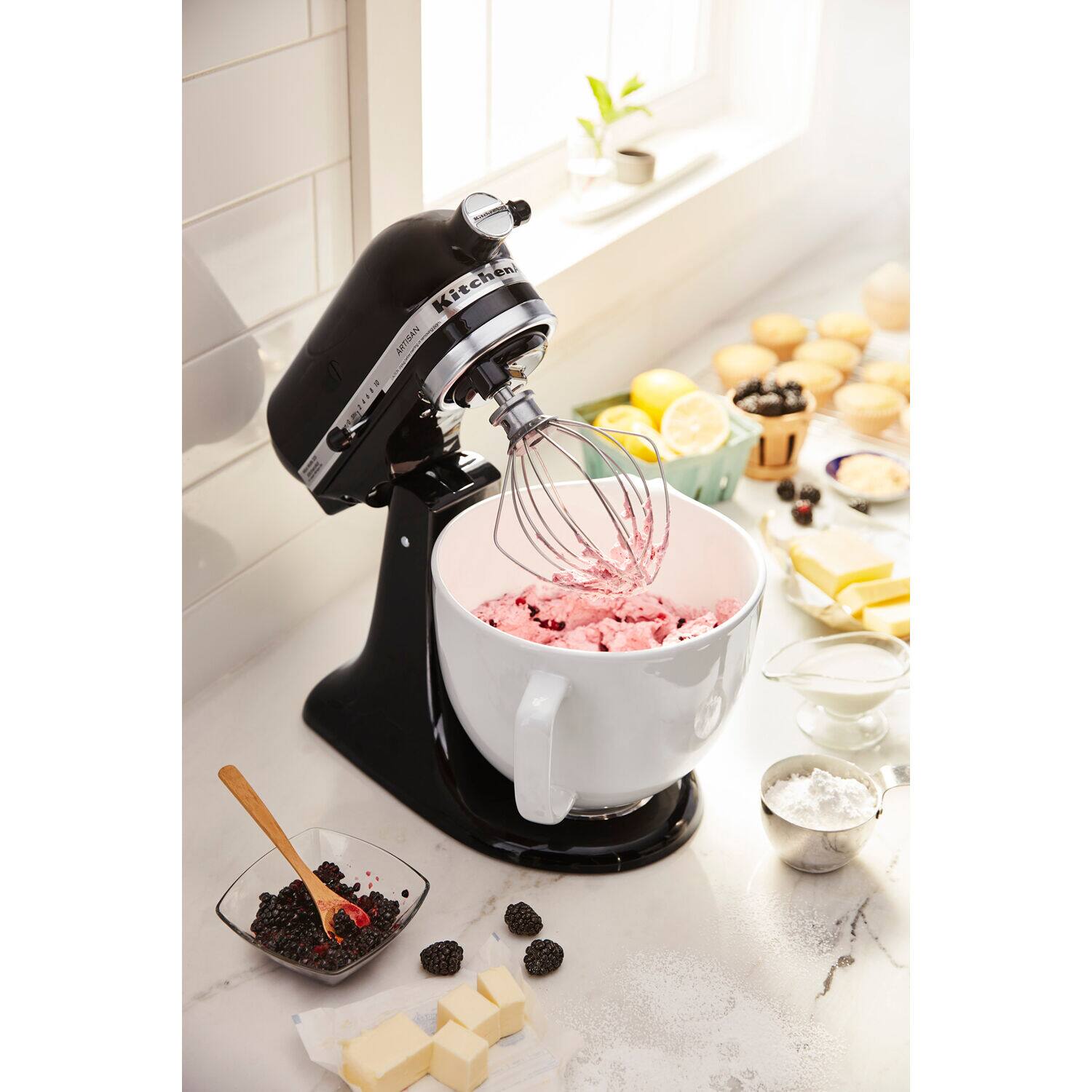 The image shows a kitchen counter with a mixer and a bowl of pink frosting. The mixer is in the process of mixing the frosting, and there are various ingredients and utensils nearby. A spoon is placed next to the bowl, and a cup can be seen in the background. The counter also has a few other bowls and cups, indicating that this is a well-equipped kitchen for baking and cooking.