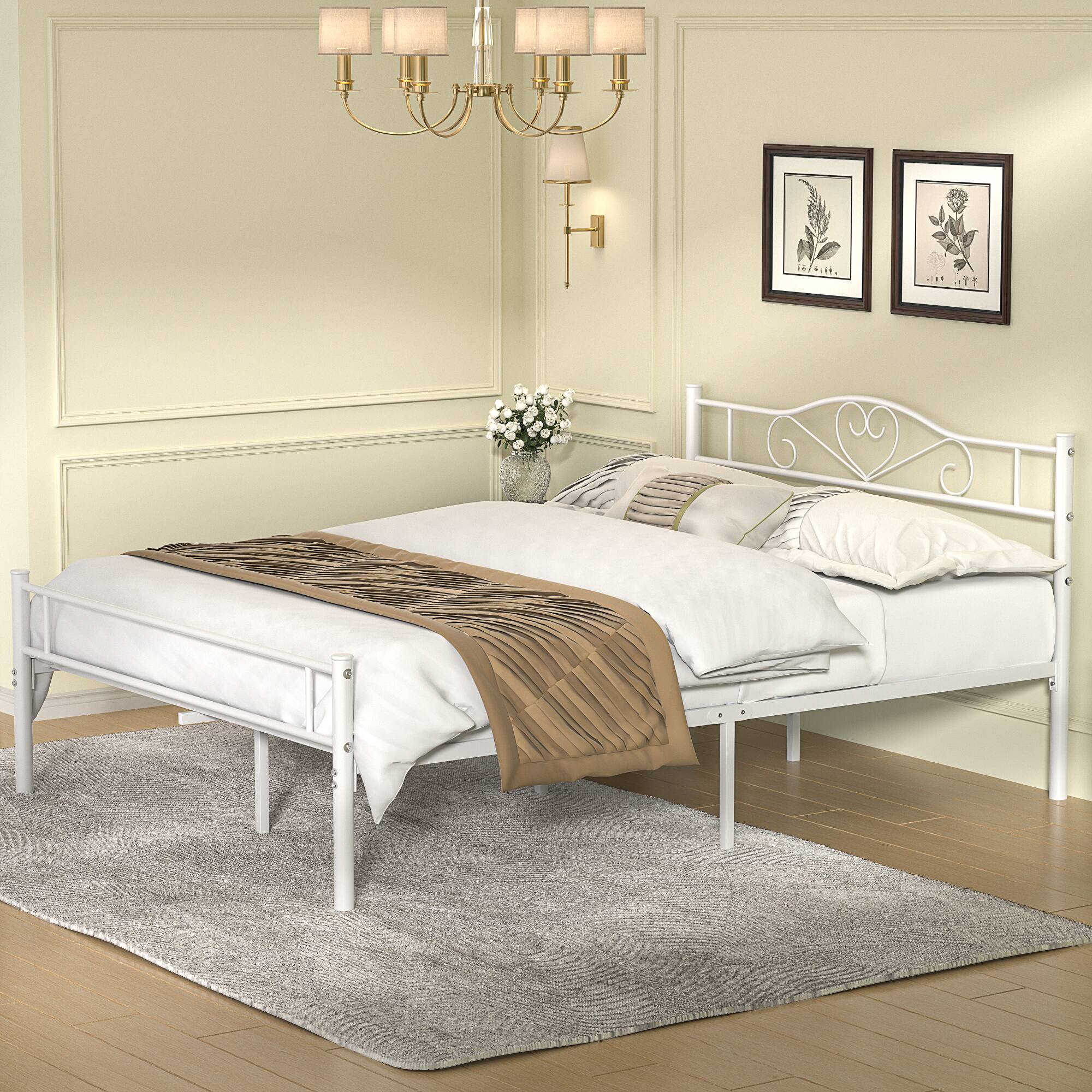 The image does not contain any visible text to group or correct. It shows a bedroom with a white metal bed, a beige and brown throw blanket, white pillows, a floral arrangement, framed botanical prints on the wall, and a chandelier.