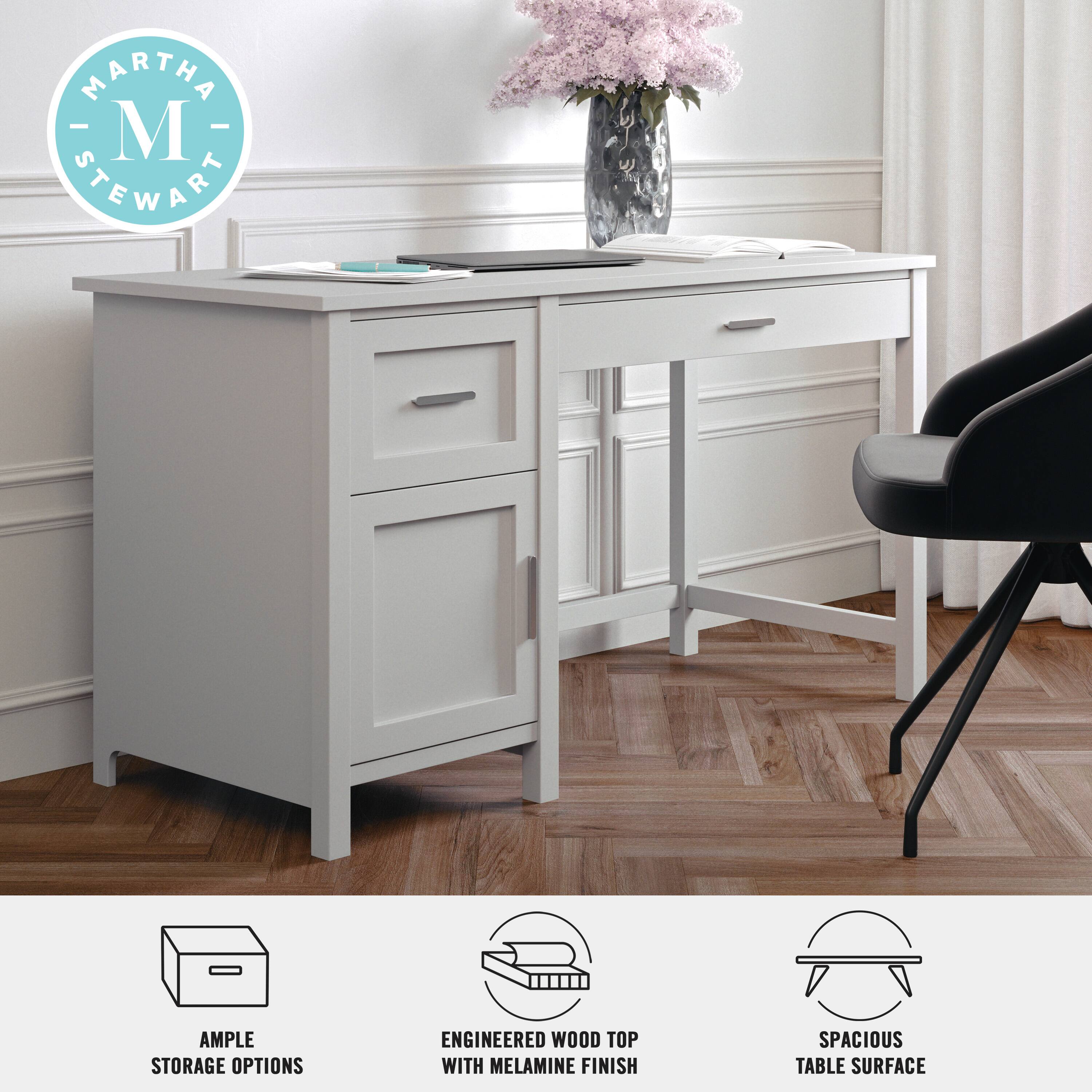 The image features a white desk with a black chair, a vase of flowers, and a pink background. The desk is designed by Martha Stewart and offers ample storage options, including an engineered wood top with a melamine finish. The desk is spacious, providing a comfortable workspace.