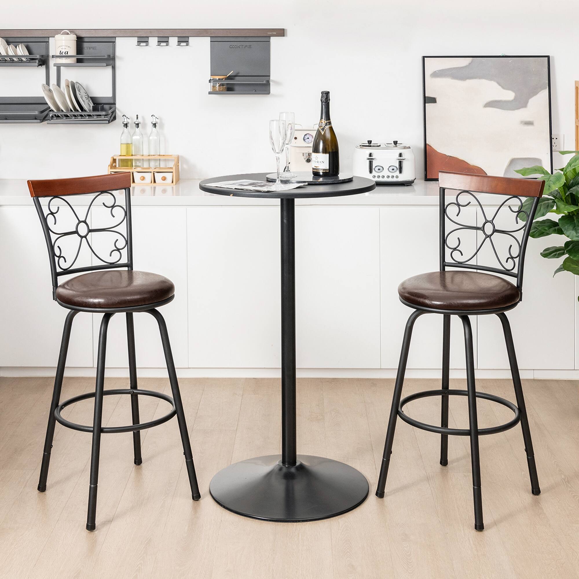 The image shows a modern kitchen or bar area with a round black table and two matching bar stools with brown leather seats. On the table, there is a bottle of champagne and two glasses. The background features a white countertop with shelves holding plates, bottles, and a small toaster. A painting and a plant are also visible.