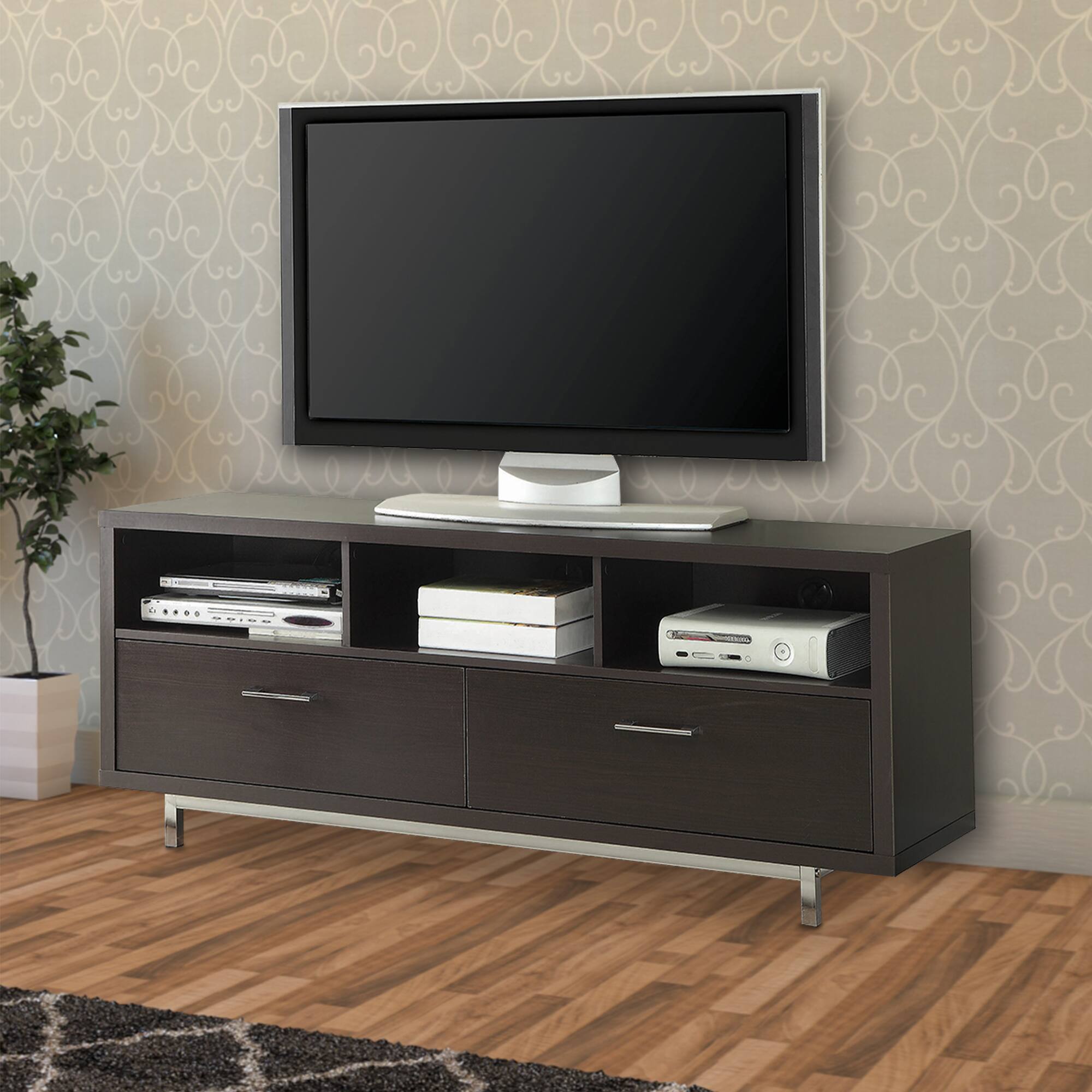 The image shows a modern entertainment unit with a flat-screen TV mounted on the wall above it. The unit has a dark wood finish with two drawers and two open shelves. On the shelves, there are various electronic devices, including a DVD player, a gaming console, and a sound system. The room has a light-colored wallpaper with a subtle pattern, a potted plant to the left, and a wooden floor with a rug in front of the unit.