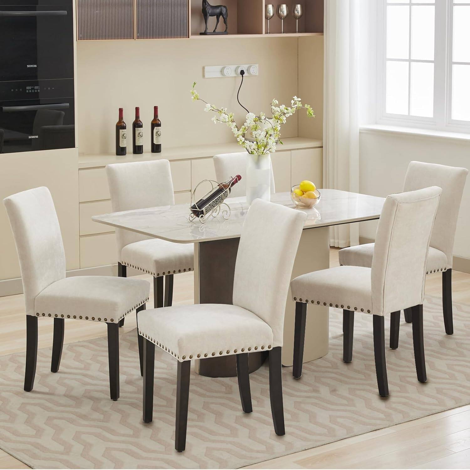 The image does not contain any text to group or correct. It shows a modern dining area with a marble table, white upholstered chairs, and a kitchen in the background. There are wine bottles and a vase with flowers on the table.