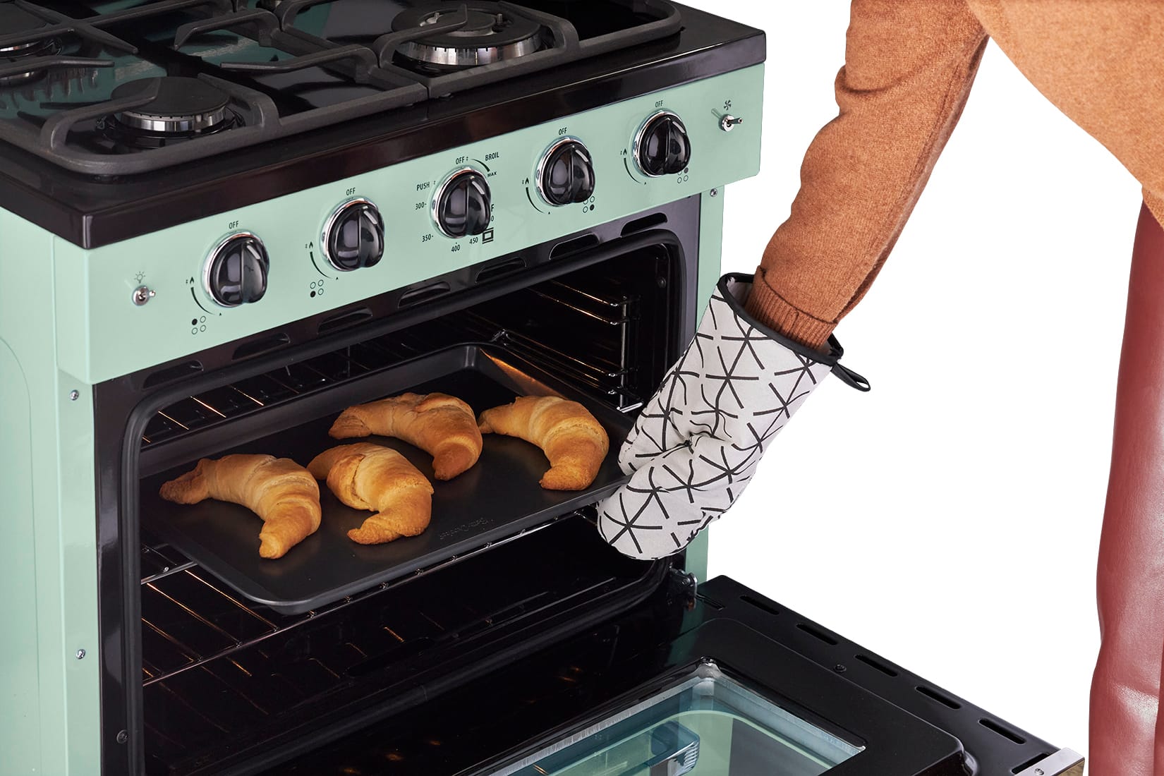 The image shows a person wearing a brown jacket and using oven mitts to take a tray of croissants out of an oven. The oven is green and has a glass door. The person is reaching into the oven to remove the tray of pastries, which includes a total of 12 croissants. The oven has a stove top with four burners, and the person is using the oven mitts to protect their hands from the heat while handling the tray.