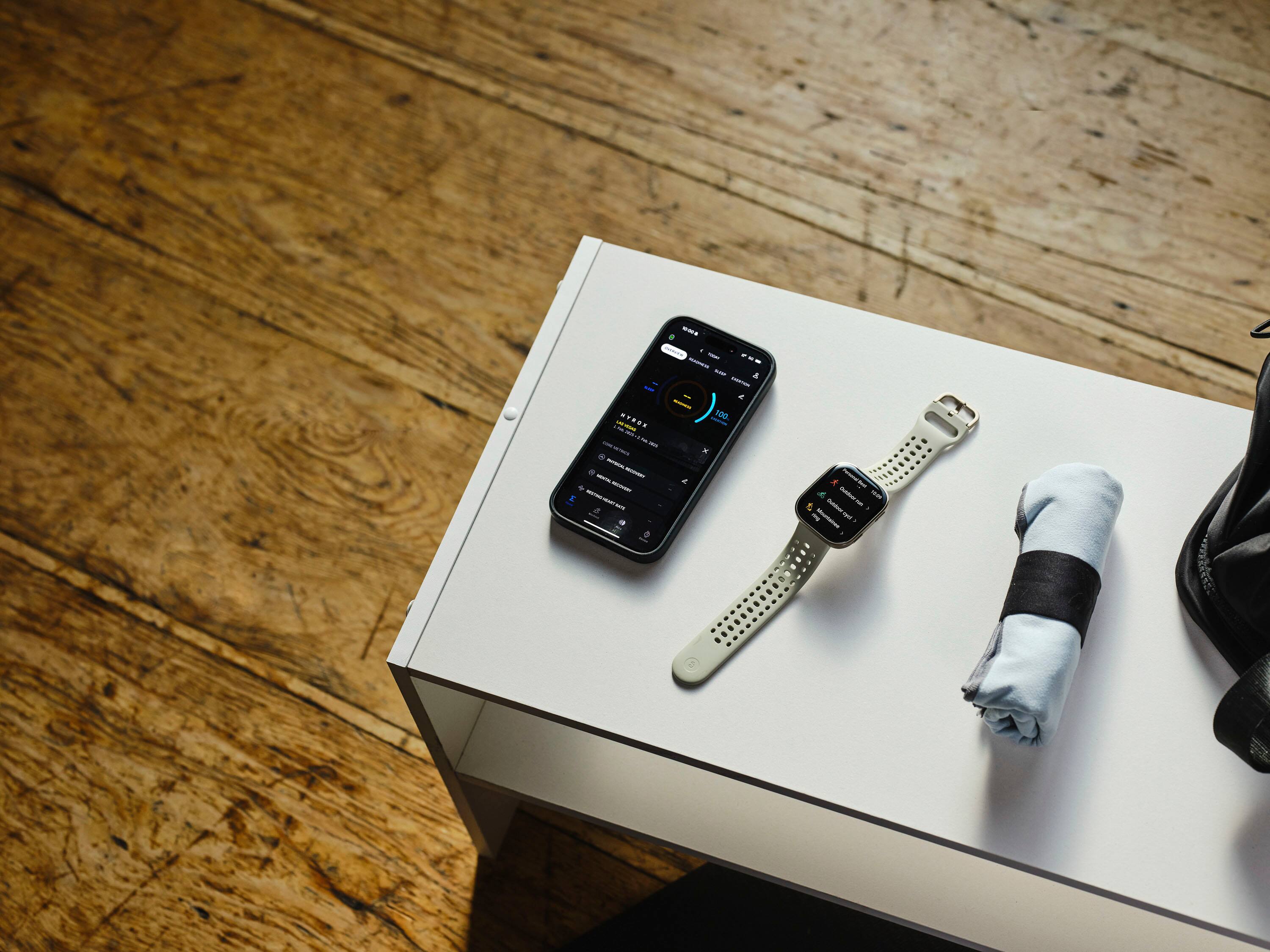 The image features a white table with a cell phone, a watch, and a towel on it. The cell phone is placed next to the watch, and the towel is located towards the right side of the table. The table is situated on a wooden floor, and the scene appears to be in a room.