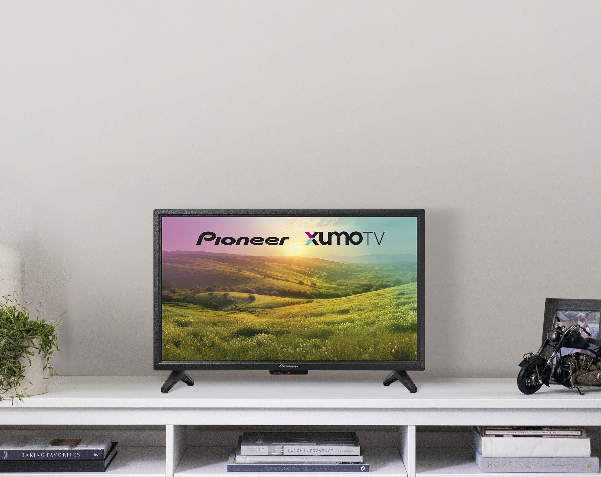 Pioneer XUMO TV is displayed on a flat screen TV. The TV is placed on a white shelf, surrounded by books and a potted plant. The TV is turned on, showcasing the Pioneer XUMO TV logo.