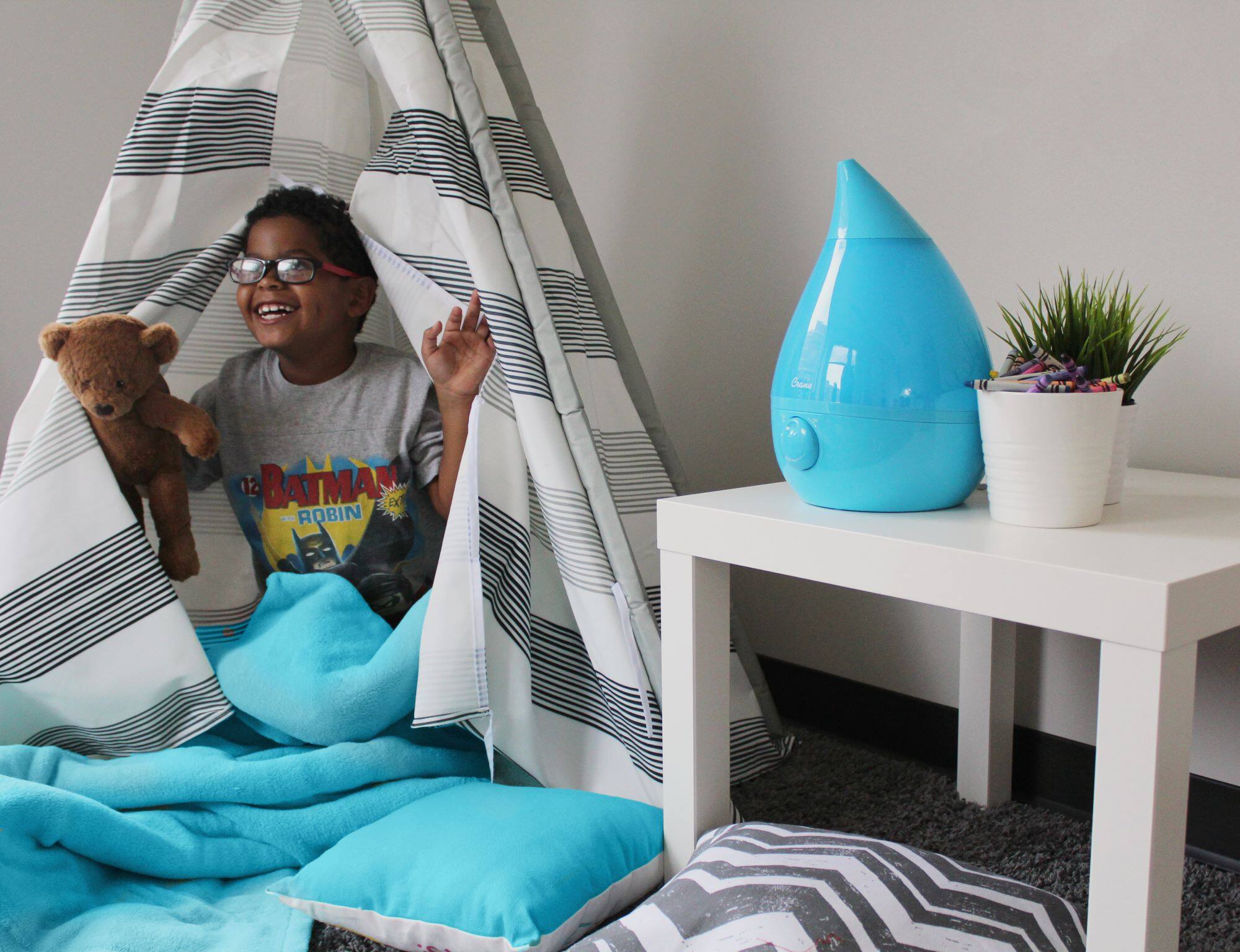 The image features a young boy sitting on a bed, holding a teddy bear. The bed is covered with a blue blanket, and there is a potted plant nearby. The boy appears to be enjoying his time with the teddy bear, possibly playing or cuddling with it. The scene captures a moment of childhood and comfort.
