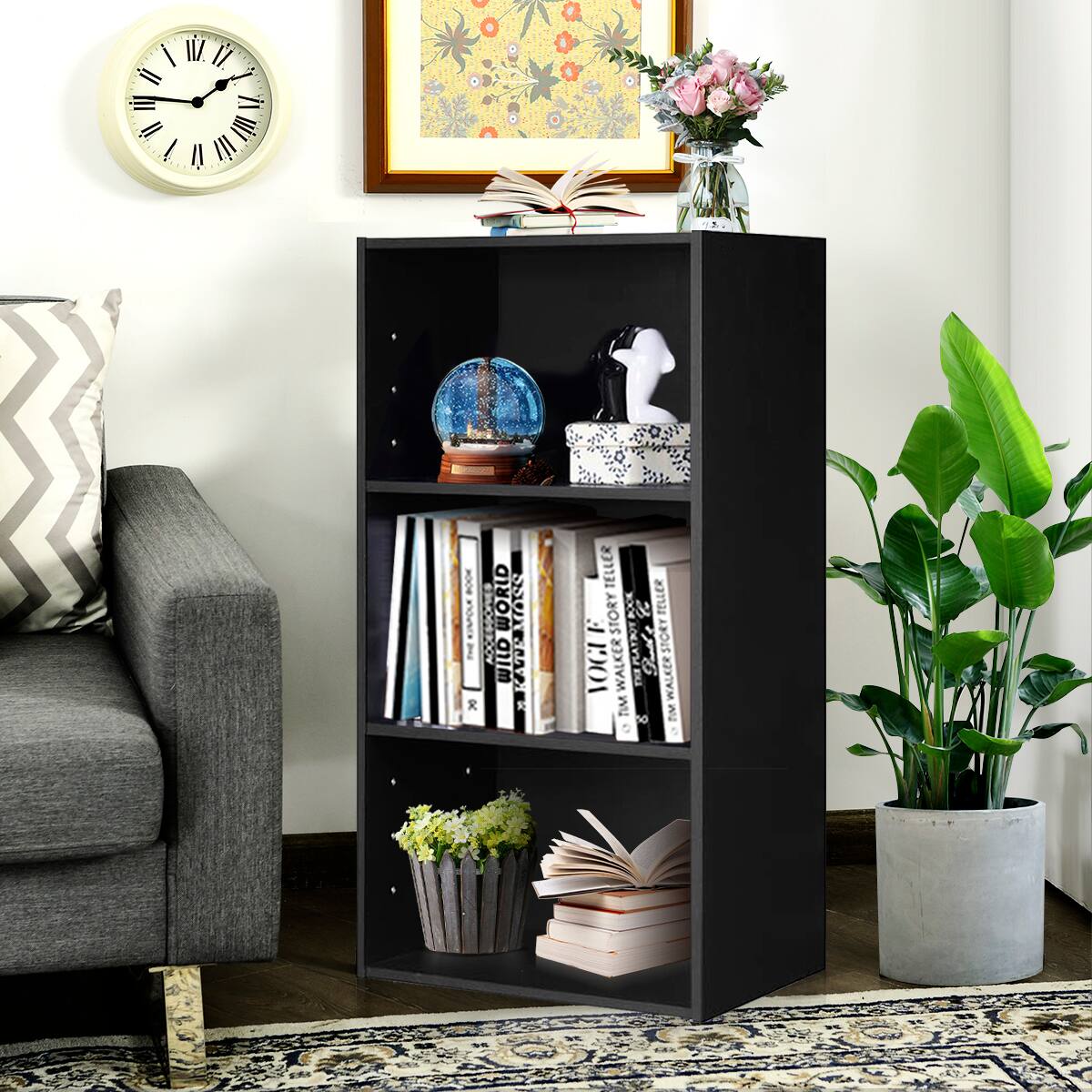 The image features a black bookshelf with a variety of books on it. The shelf is located in a living room, with a couch in the background. The bookshelf is filled with books, and there is also a clock on the wall above the bookshelf. Additionally, there is a vase with flowers on the bookshelf, adding a touch of decoration to the space.