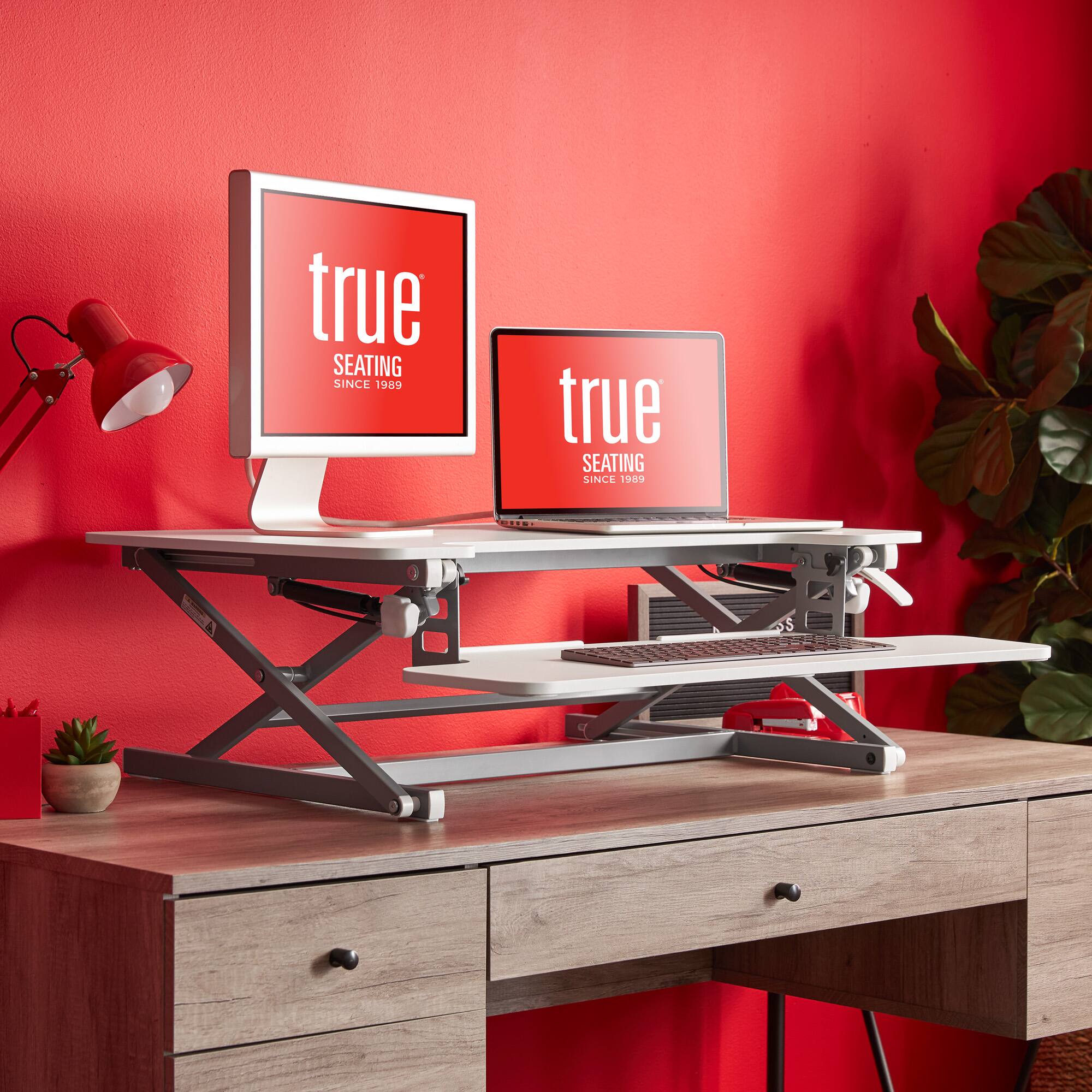 The image features a desk with a laptop and a monitor on it. The laptop is placed on a stand, and there is a red wall behind the desk. The desk is positioned in front of a red wall, which adds a vibrant touch to the workspace. The desk setup includes a keyboard and a mouse, providing a comfortable and functional workspace.