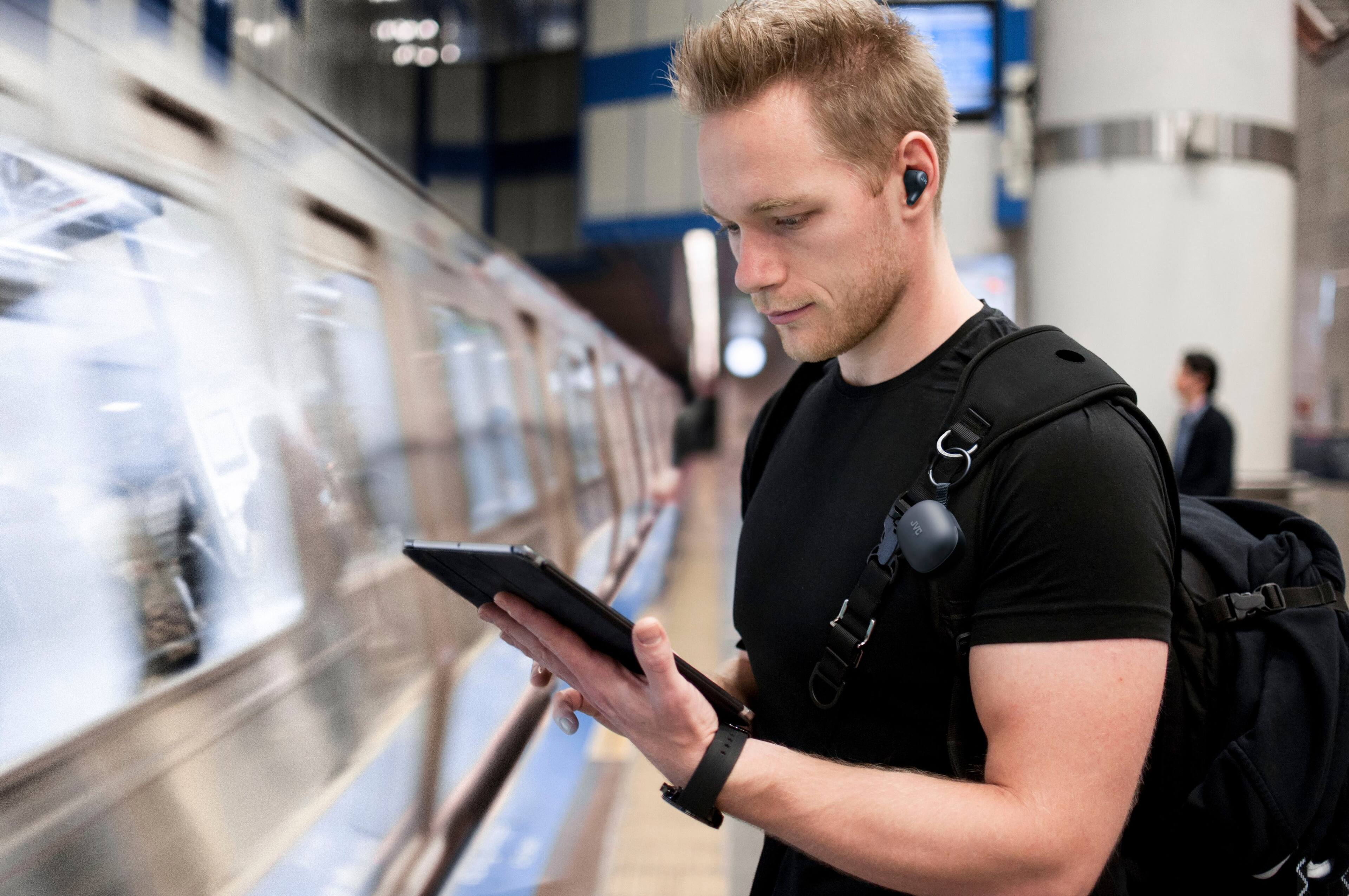 The image shows a man standing in a subway train, holding a tablet and looking at it. He is wearing a black shirt and has a backpack on. The man is also wearing headphones, possibly listening to music or an audiobook. The subway train is moving, and the man seems to be focused on his tablet, possibly checking the train schedule or using a navigation app.