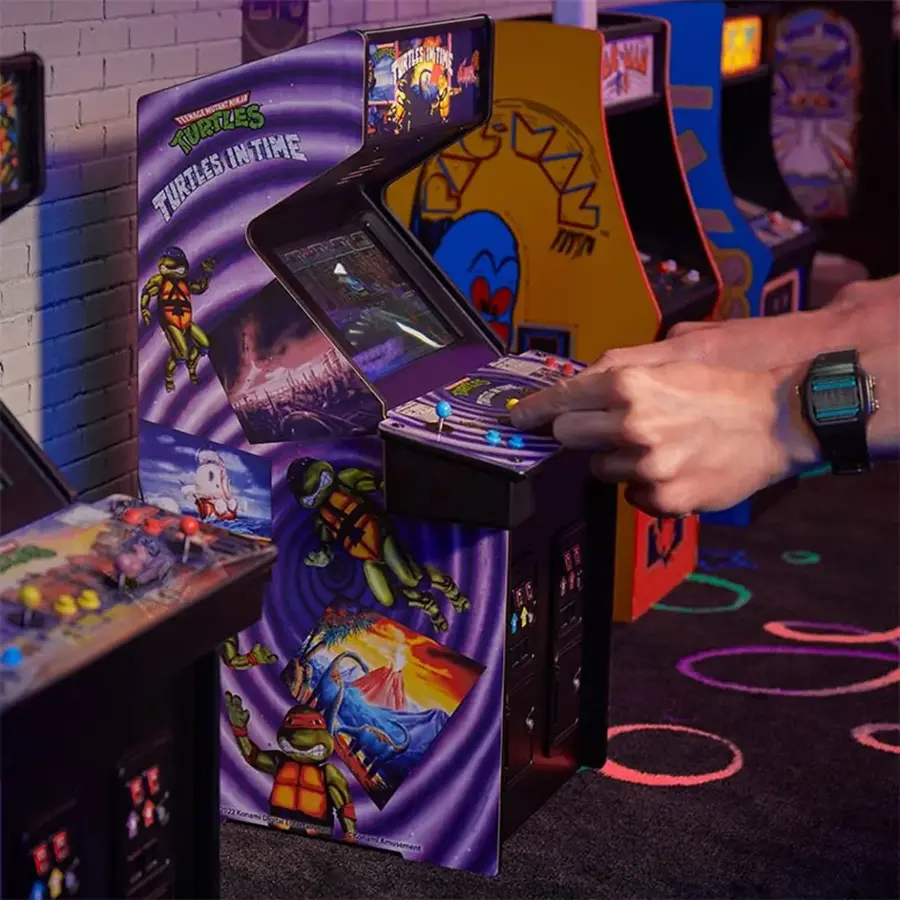 The image features a person playing a video game, specifically Teenage Mutant Ninja Turtles in Time, on a Nintendo Gameboy. The game is displayed on a TV screen, and the person is using a remote control to play. The scene also includes a clock on the wall, and there are several other video game machines in the background.