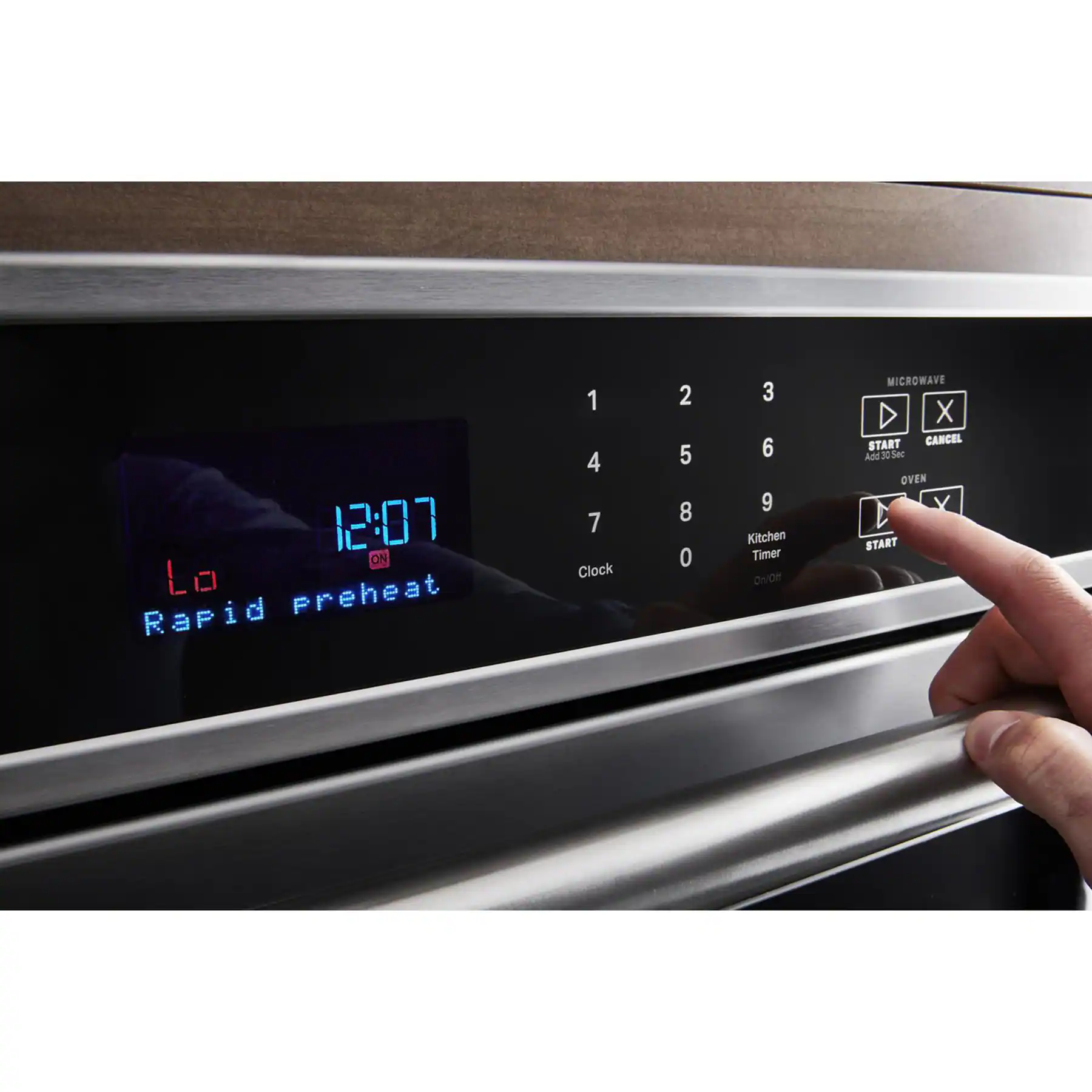 The image shows a microwave with a timer set to 12:07. A person is pointing at the timer, which is set to "rapid preheat." The timer also has buttons for "1," "4," "7," "Clock," "2," "5," "8," "0," "3," "6," and "9." The microwave also has a "Kitchen Timer" button and a "Start" button.