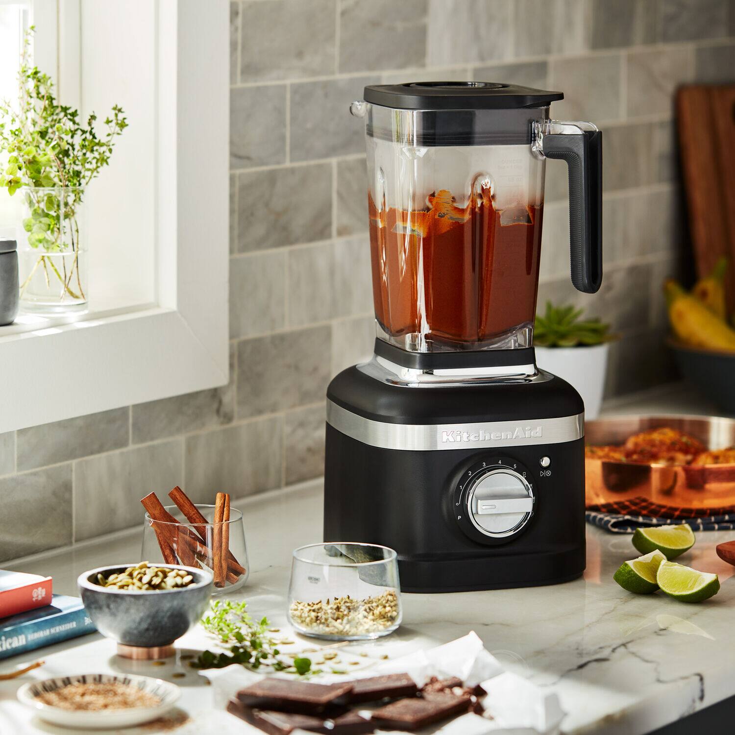 KitchenAid is a brand of blenders and other kitchen appliances. The image shows a KitchenAid blender sitting on a counter, surrounded by various ingredients and food items.
