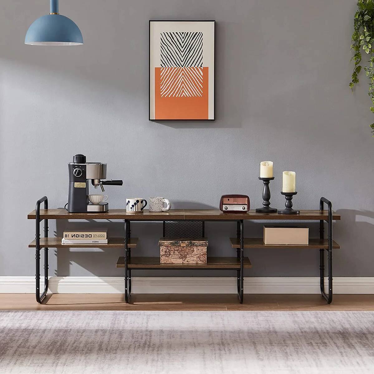 The image shows a minimalist interior design featuring a wooden console table with black metal legs. On the table, there is a black coffee machine, a white mug, a small white teapot, and a few other items. Above the table, a framed abstract art piece with geometric patterns in black and orange is hung on the wall. To the right, there are two candles in black holders and a small vintage-style radio. The table also has a few decorative items, including a book titled "18 DESIGN" and a map in a wooden frame. The floor is covered with a light-colored rug, and a blue pendant light hangs from the ceiling.
