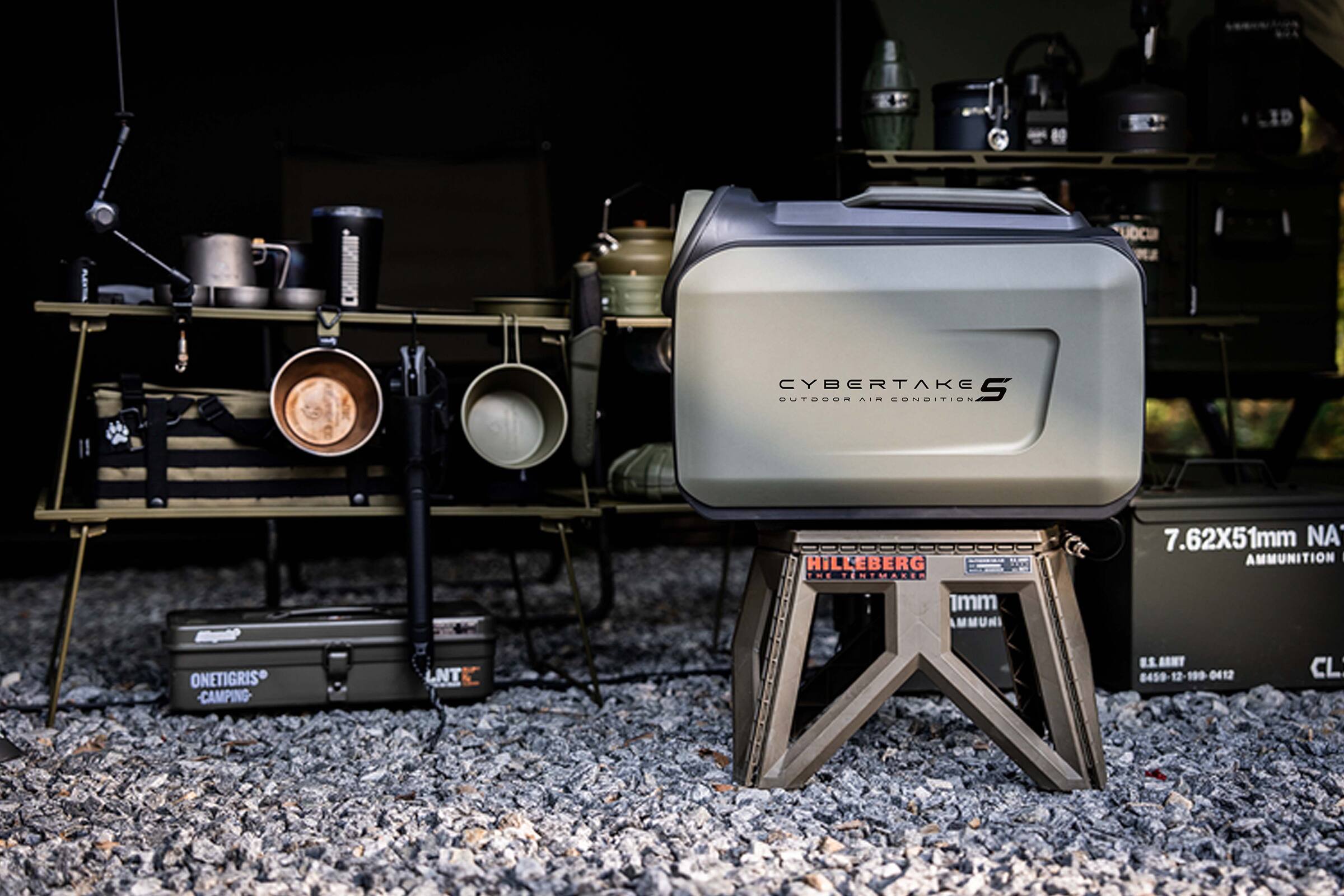 The image features a green Cybertake S camera on a stand, surrounded by various camping equipment. The stand is placed on a gravel surface, and there are several other items in the scene, including a backpack, a bottle, and a cup. The presence of these items suggests that the location is a campsite, and the camera is being used for outdoor photography or filming.