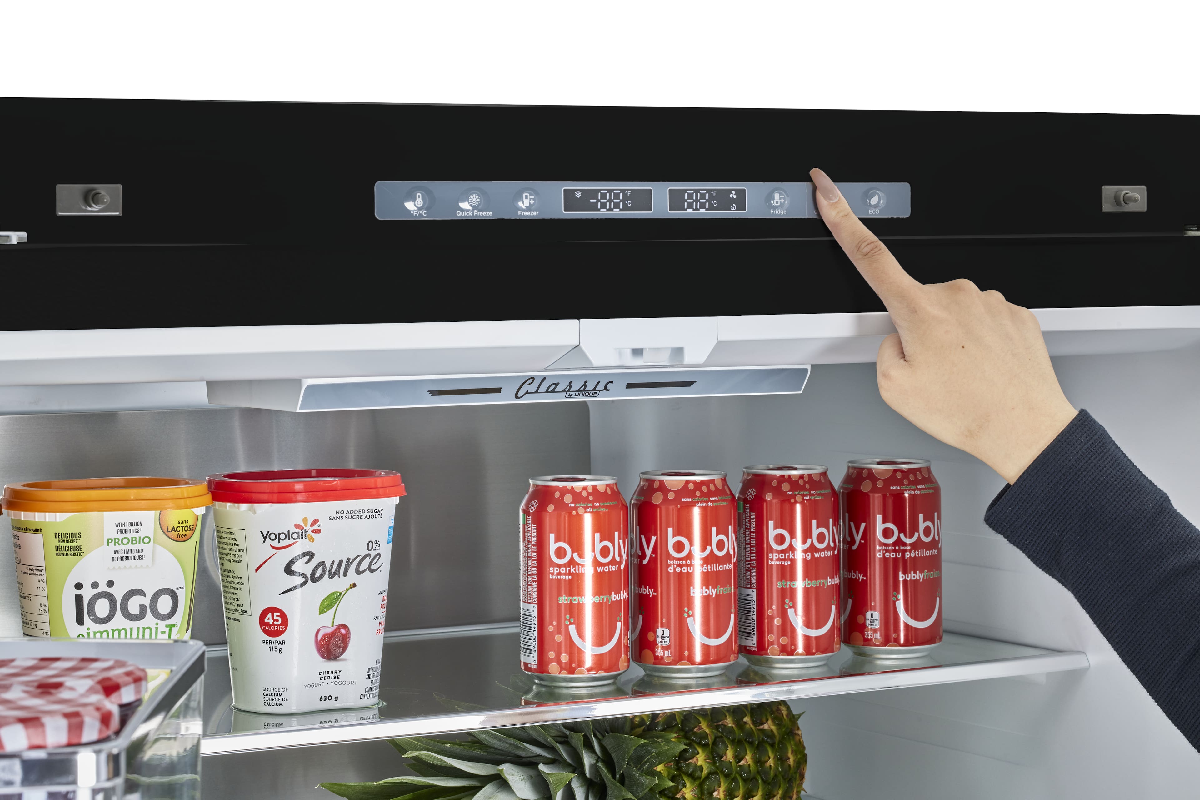 The image shows a person pointing at a refrigerator with a variety of drinks inside. The drinks include a bottle of strawberry bubly, a bottle of iced tea, and several cans of soda. The person is likely selecting a drink to enjoy.