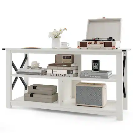 A white table with a vase of flowers, a coffee mug, a suitcase, and a bookshelf with various books and magazines. The table also has a pair of speakers, a cup, and a bowl on it.