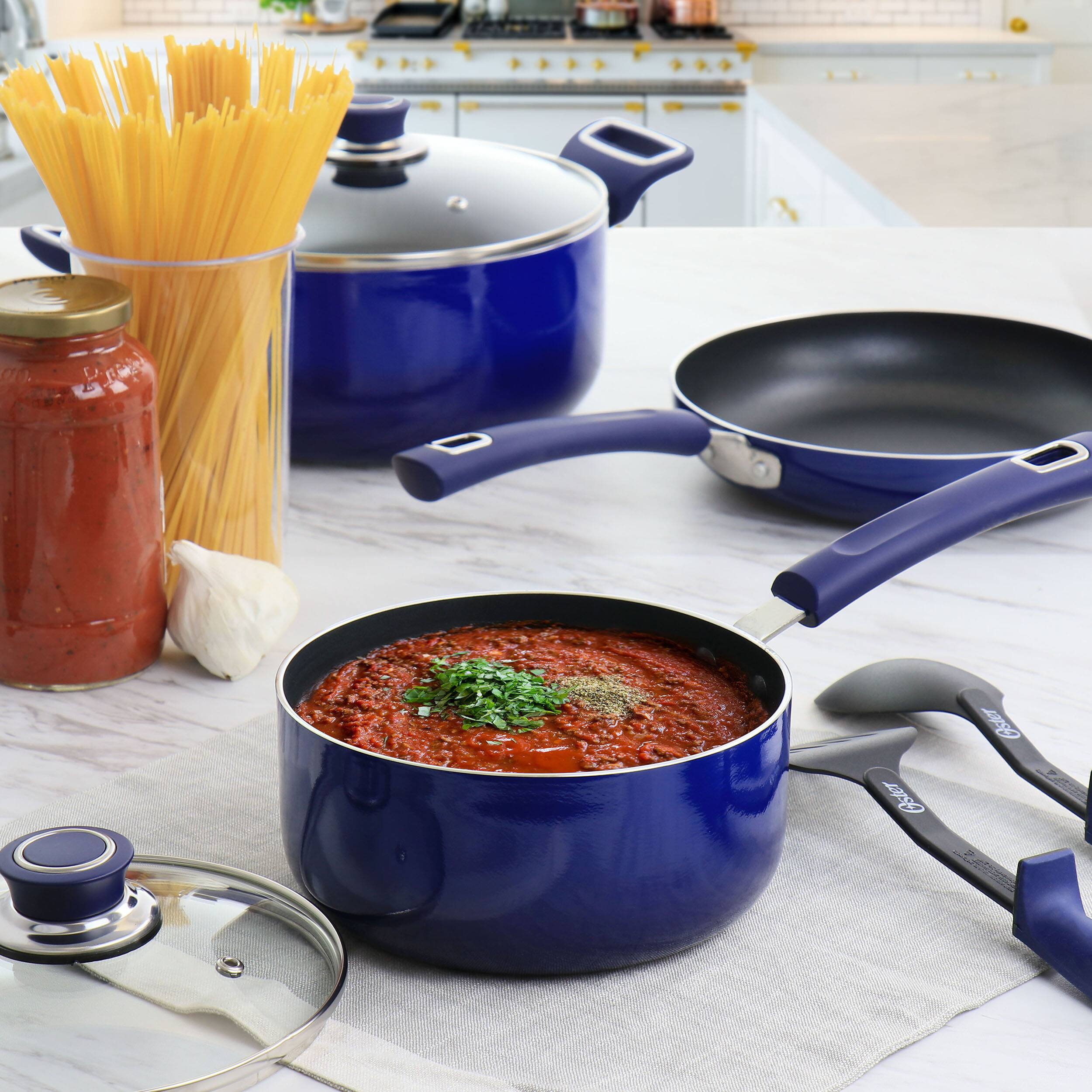The image features a blue pot filled with red sauce, placed on a white countertop. The pot is accompanied by a blue pan and a blue lid, both of which are also filled with red sauce. The countertop also has a blue bowl and a blue spoon, both of which are placed close to the pot. Additionally, there is a jar of red sauce on the countertop, and a spaghetti noodle is visible near the pot.