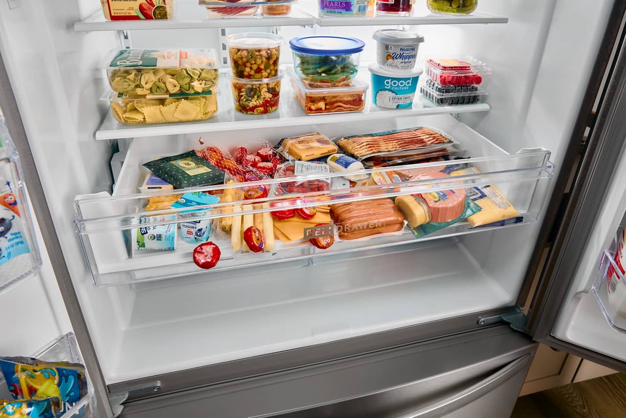 The refrigerator is filled with various food items. The top shelf contains several bottles, while the middle shelf has a variety of foods such as cheese, tomatoes, and hot dogs. The bottom shelf has a drawer with more food items, including yogurt. The refrigerator is well-stocked, providing a variety of options for meals and snacks.