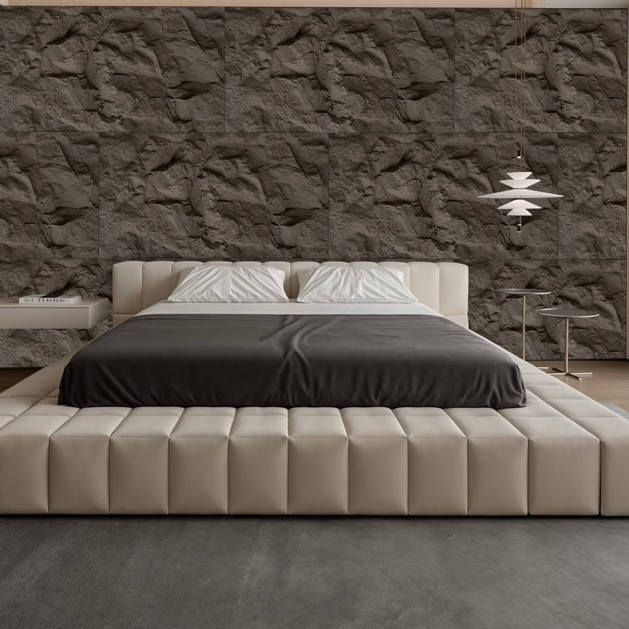 The image shows a modern bedroom with a large bed featuring a beige upholstered headboard and a dark gray bedspread. The wall behind the bed has a textured, dark stone finish. There is a small bedside table with a book on it, and a modern pendant light hanging from the ceiling. The floor is a dark, smooth surface.