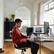 The man is sitting at a desk with a computer monitor, a keyboard, and a mouse. He is wearing a red shirt and is looking at the screen. There is a cup on the desk, and a potted plant is nearby. The man is also holding a book, possibly reading or studying.