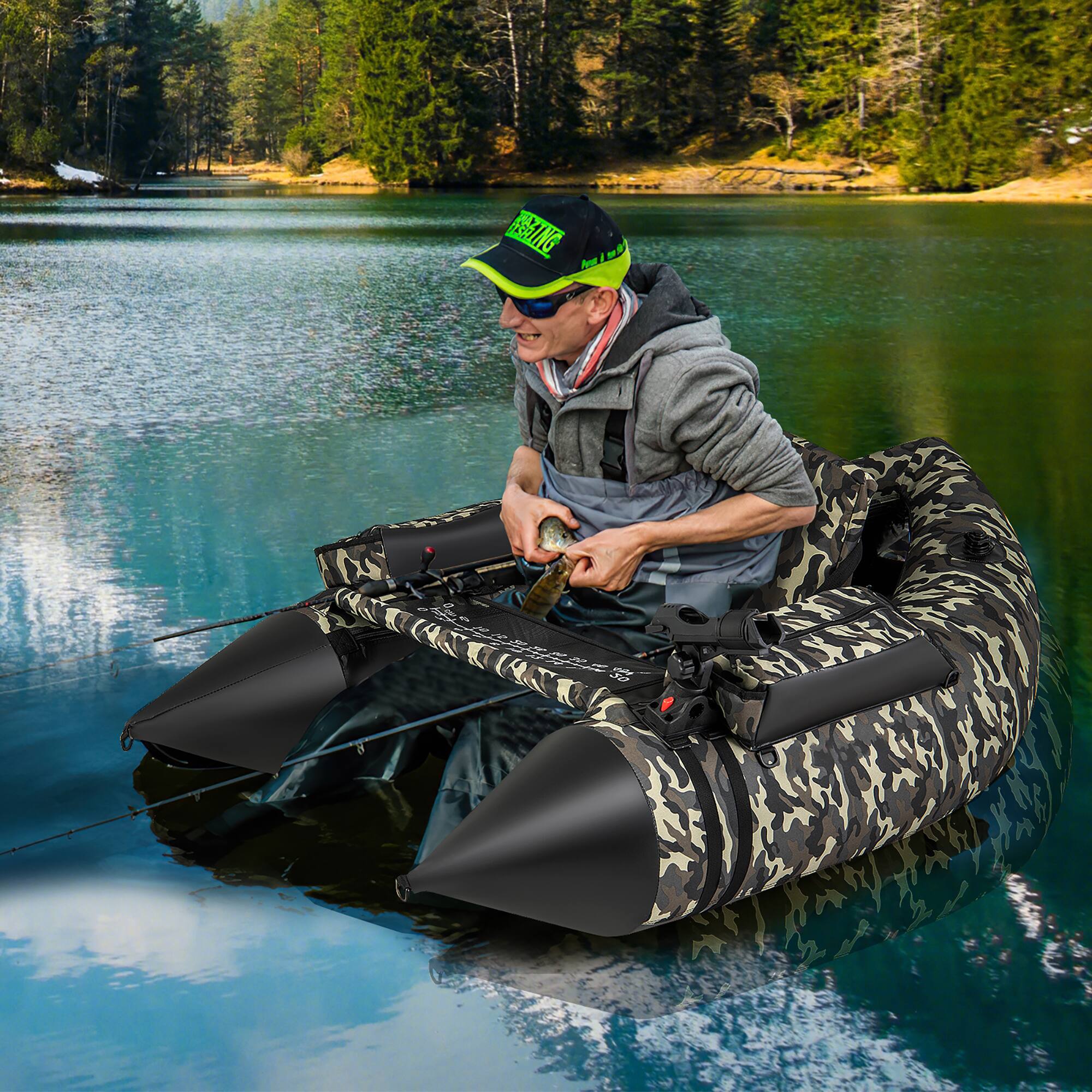 A man is sitting on an inflatable boat in a lake, holding a fish. He is wearing a cap and a jacket. The boat has a camouflage pattern.