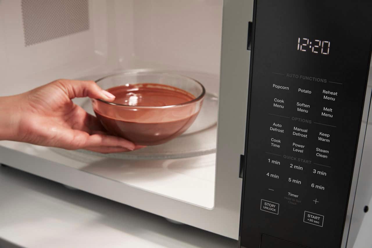The image shows a microwave with a bowl of chocolate inside it. The microwave is set to 12:20, and the timer is set to 3 minutes. The menu options include auto functions such as popcorn, potato reheat, cook, and melt. There are also manual defrost and keep cook warm options available. The power level can be set to various levels, and the timer can be set to different intervals, including 1 minute, 2 minutes, 3 minutes, 4 minutes, 5 minutes, 6 minutes, and 7 minutes. The image also features a clock that can be used to set the timer.