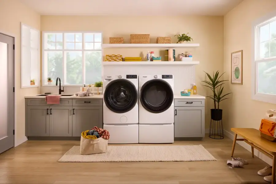 The image shows a kitchen with a washer and dryer combo in the middle of the room. The washer is white and has a black door, while the dryer is also white. The kitchen features a sink, a dishwasher, and a refrigerator. There are also several bottles placed around the kitchen, and a potted plant is located near the dryer. The room is well-lit, and the overall setting appears to be a clean and organized space.