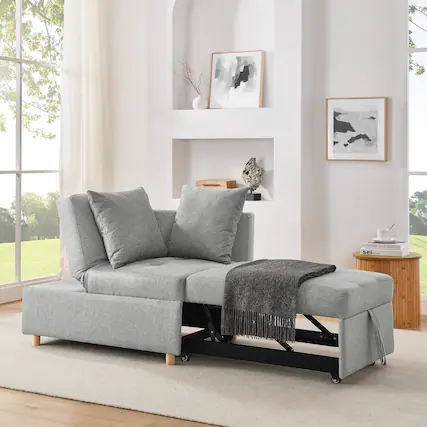 The image shows a modern living room featuring a gray fabric sofa with two cushions and a matching throw blanket draped over one arm. The sofa is positioned on a light-colored rug, with a wooden side table beside it holding a book and a cup. The room has large windows with sheer curtains, allowing natural light to fill the space. The walls are decorated with abstract art pieces and shelves holding decorative items.