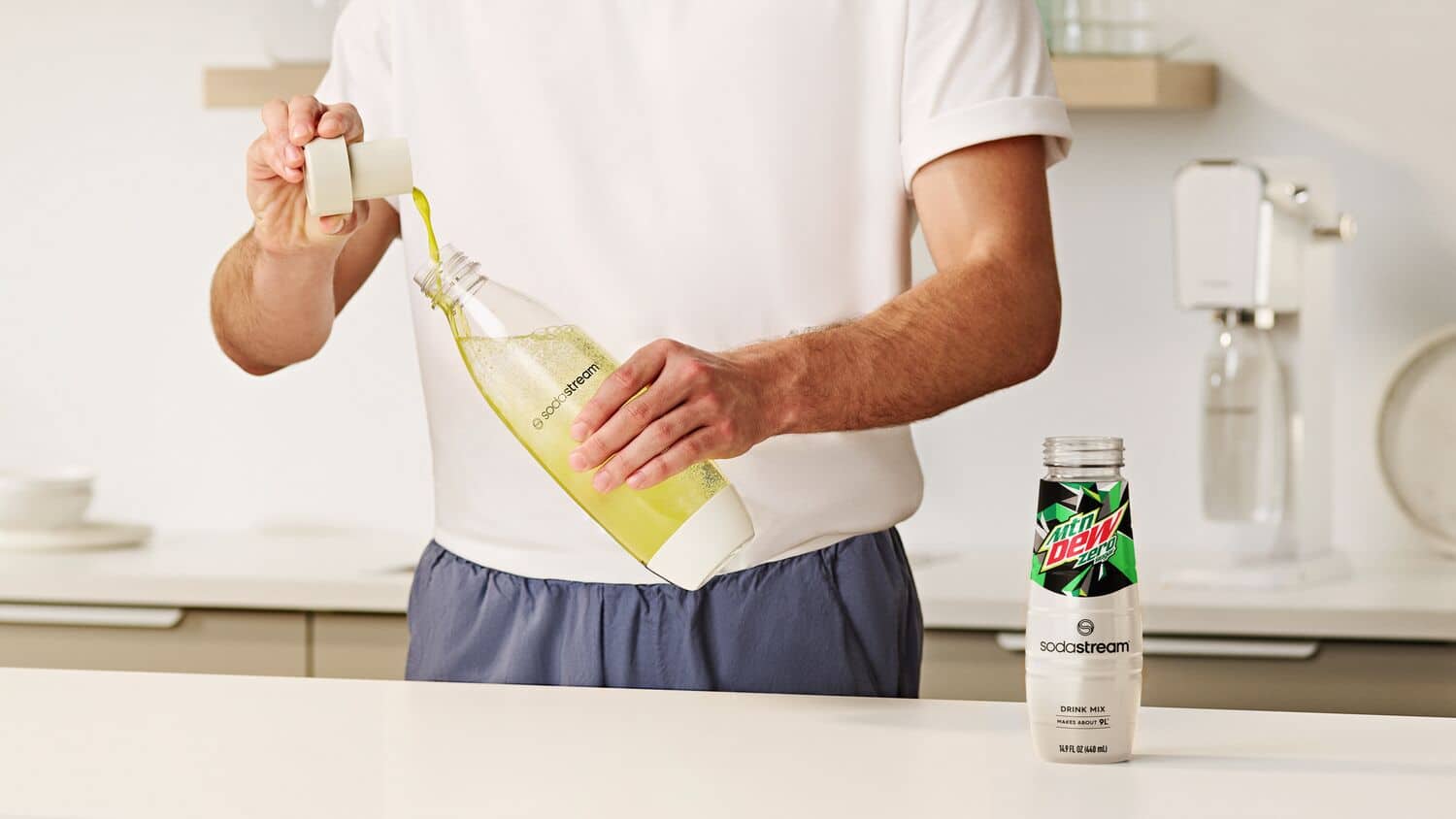 The man is holding a bottle of soda water and a can of soda mix. He is standing in a kitchen, preparing to make a drink using the soda stream.