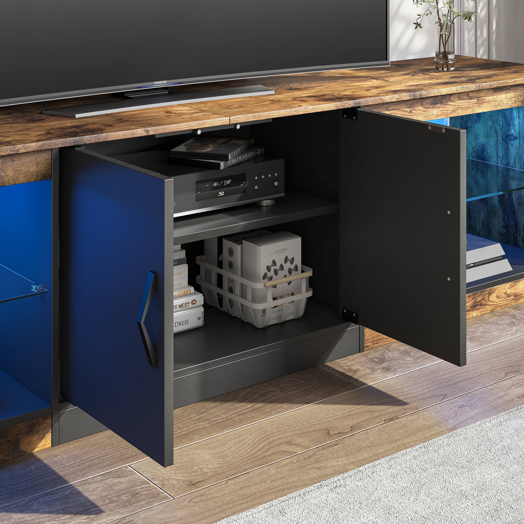 The image shows a wooden entertainment center with a television on top. The TV is placed on a wooden stand, and there is a blue drawer below it. The drawer is open, revealing a collection of items, including a remote control, a keyboard, and a mouse. There are also several books placed in the drawer. The entertainment center is situated in a living room, and a potted plant can be seen in the background.